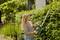 A woman is trimming a dense hedge with an electric hedge trimmer in a garden. Trees can be seen in the background.