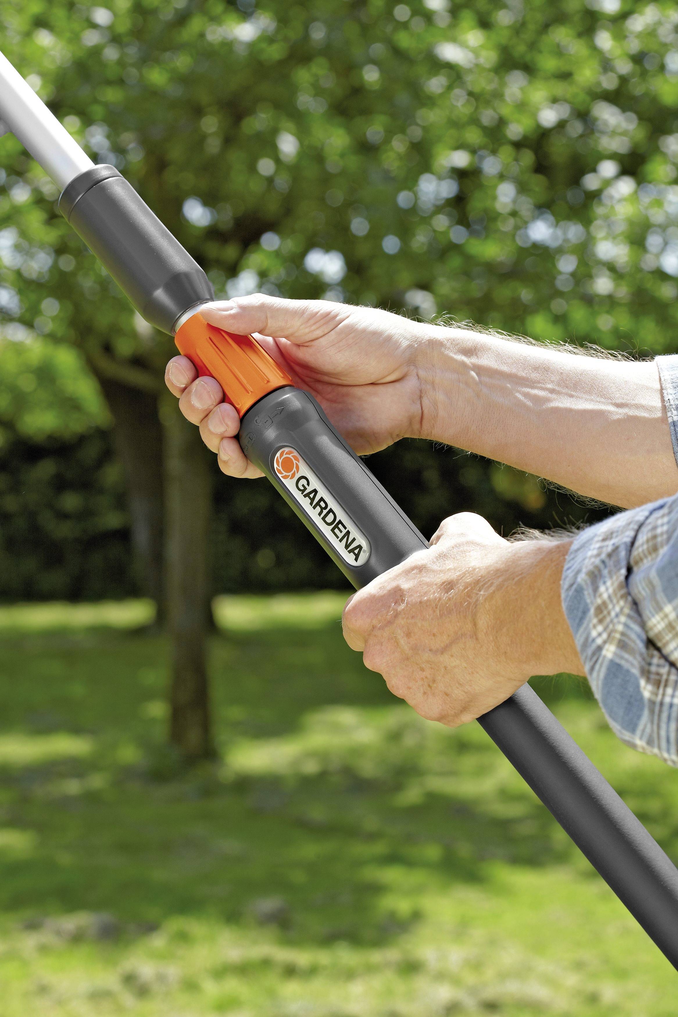 A person is holding gardening equipment with an extendable handle outdoors, with trees and lawn blurred in the background.