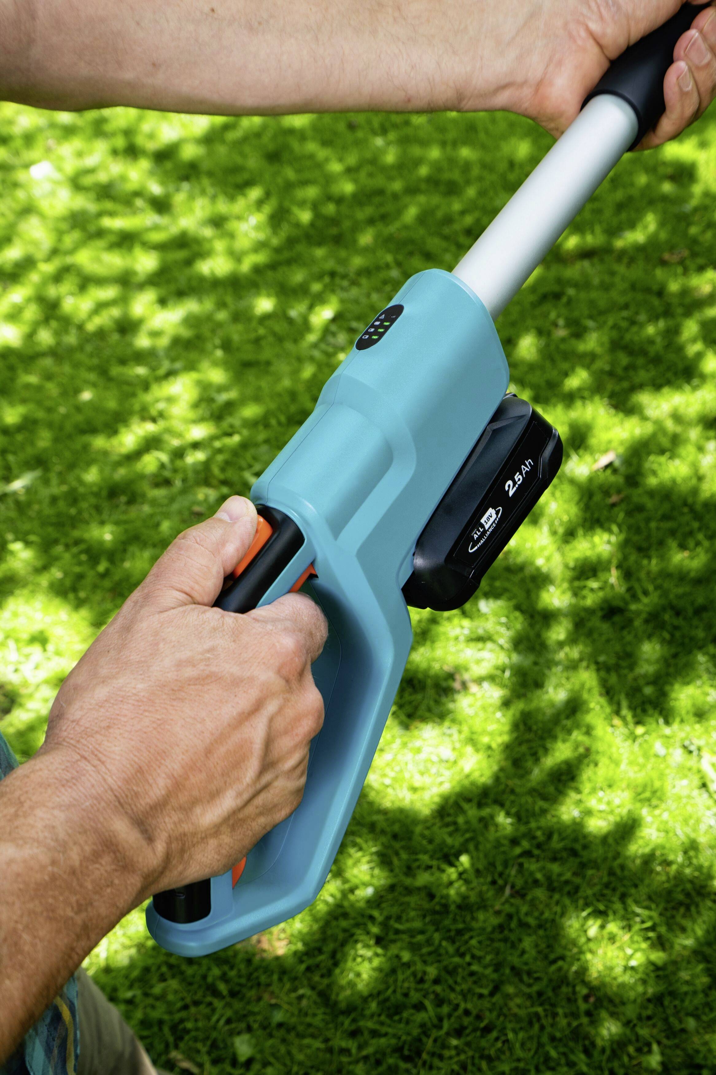 A person is holding a blue garden tool with a battery on green grass. Their hands are positioned on the handle.
