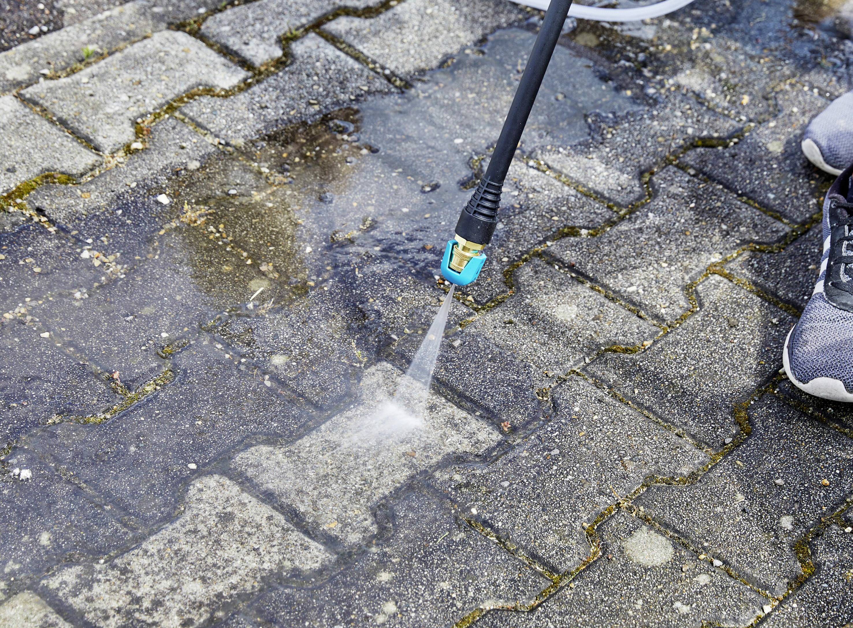 A high-pressure cleaning nozzle sprays water onto wet paving stones and removes dirt. A pair of shoes is visible at the edge.