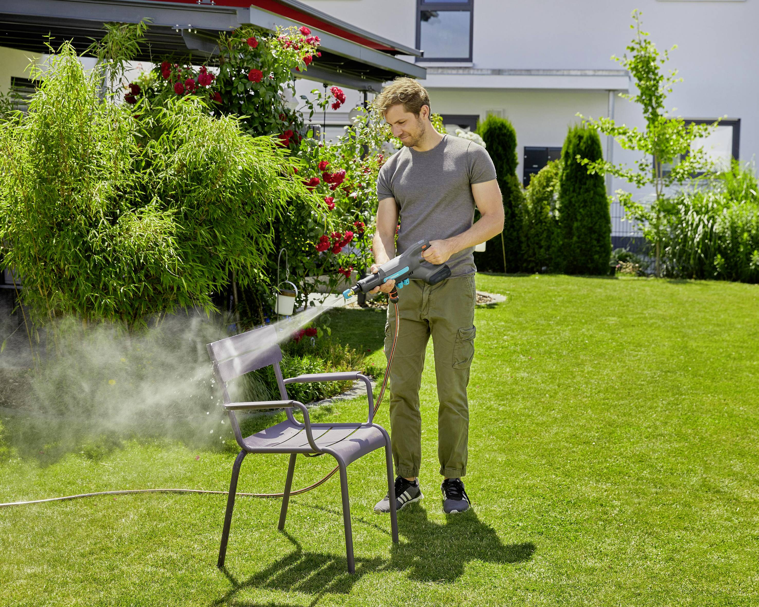 A person is cleaning a garden chair in the garden using a pressure washer. Plants and a house can be seen in the background.