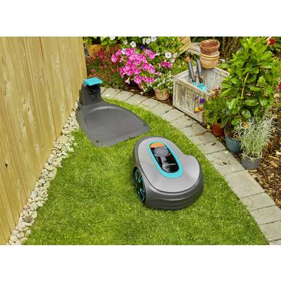 A robotic lawnmower is cutting the grass in a garden. In the background, colourful flowers are blooming and a wooden shelving unit with gardening tools stands nearby.