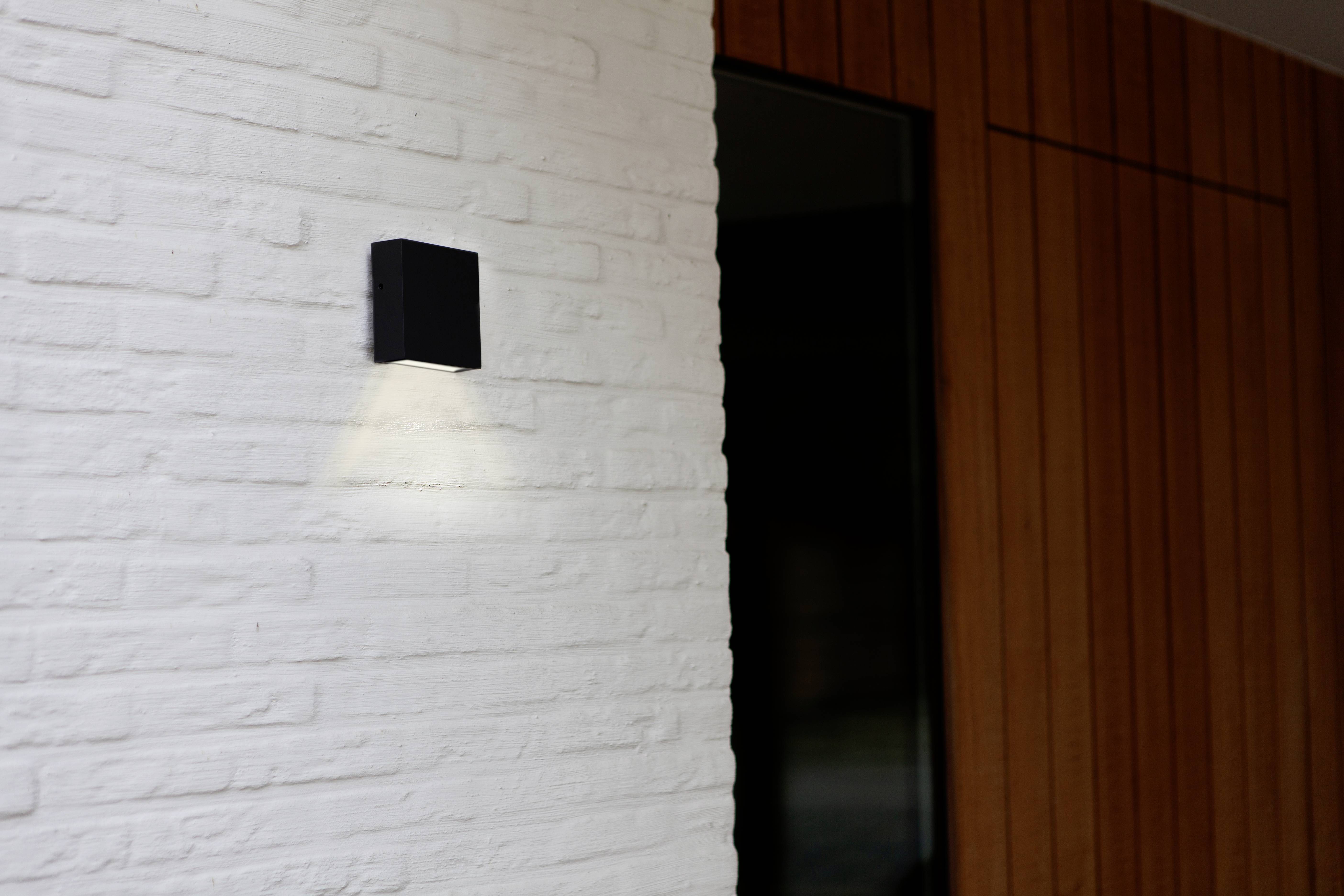 External light fitting on a white brick wall, casting a warm downward beam of light, positioned next to a wooden wall and a dark window.