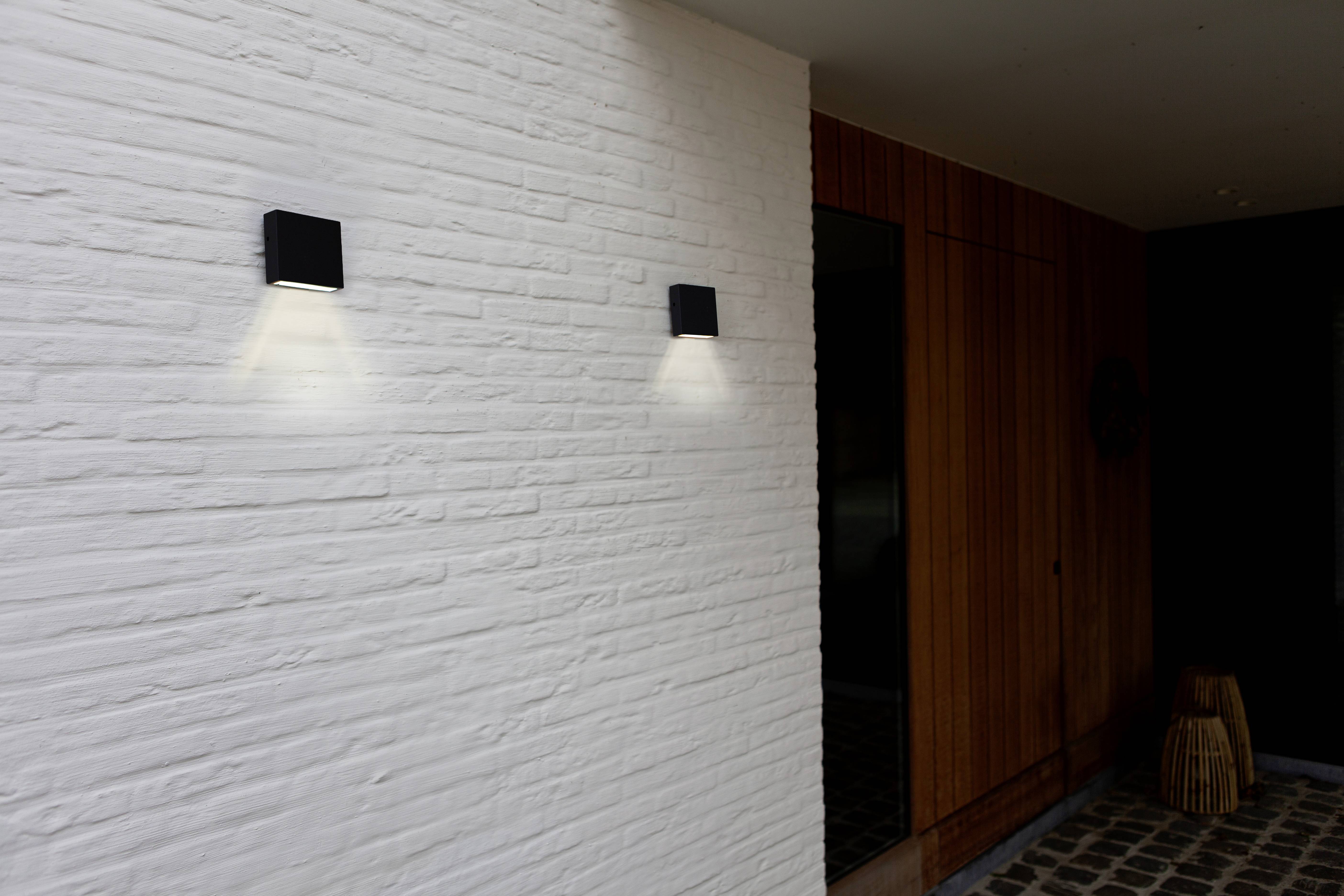 An entrance with a white brick wall and two black wall lights emitting soft light. A dark wooden door and cobblestones in the foreground.