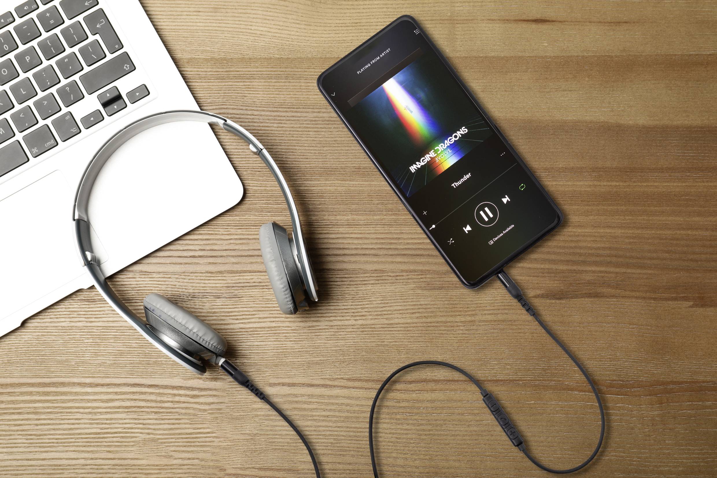 Smartphone with an open music player displaying the album 'Rainbow Roads'. Headphones connected beside a laptop on a wooden table.