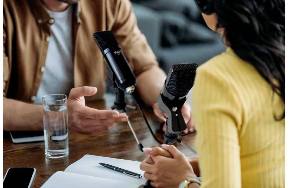 Two people are sitting at a table and chatting about microphones. A notebook, a pen, and a glass of water are lying on the table.