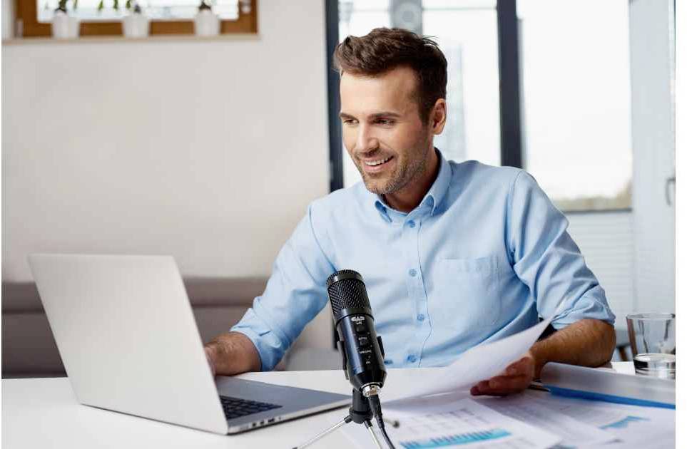 A man is sitting at a desk, working on a laptop and speaking into a microphone. Documents with graphics are spread out in front of him.
