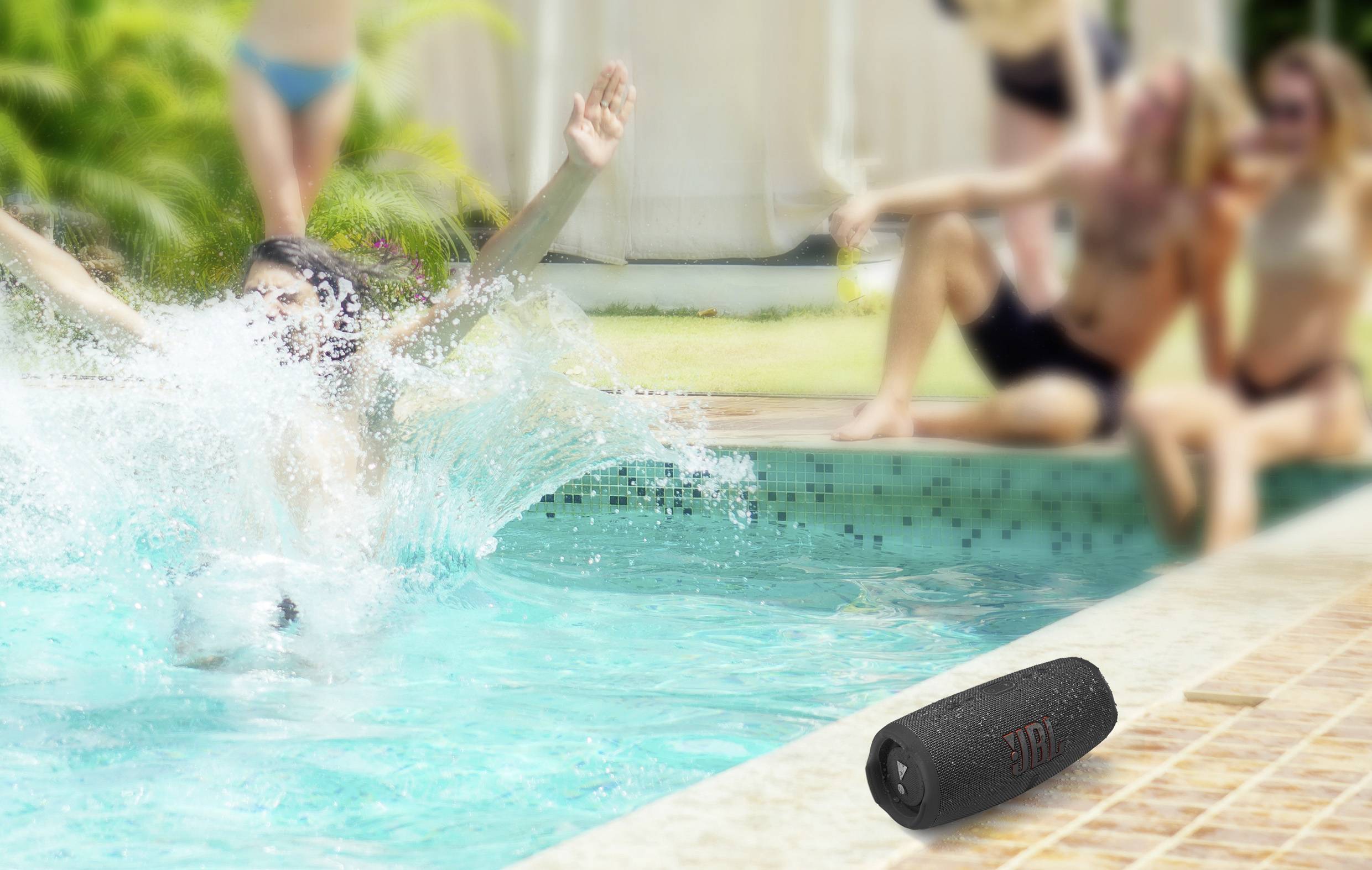 A person jumps into the pool while others sit around the edge. A black speaker lies in the foreground by the poolside.