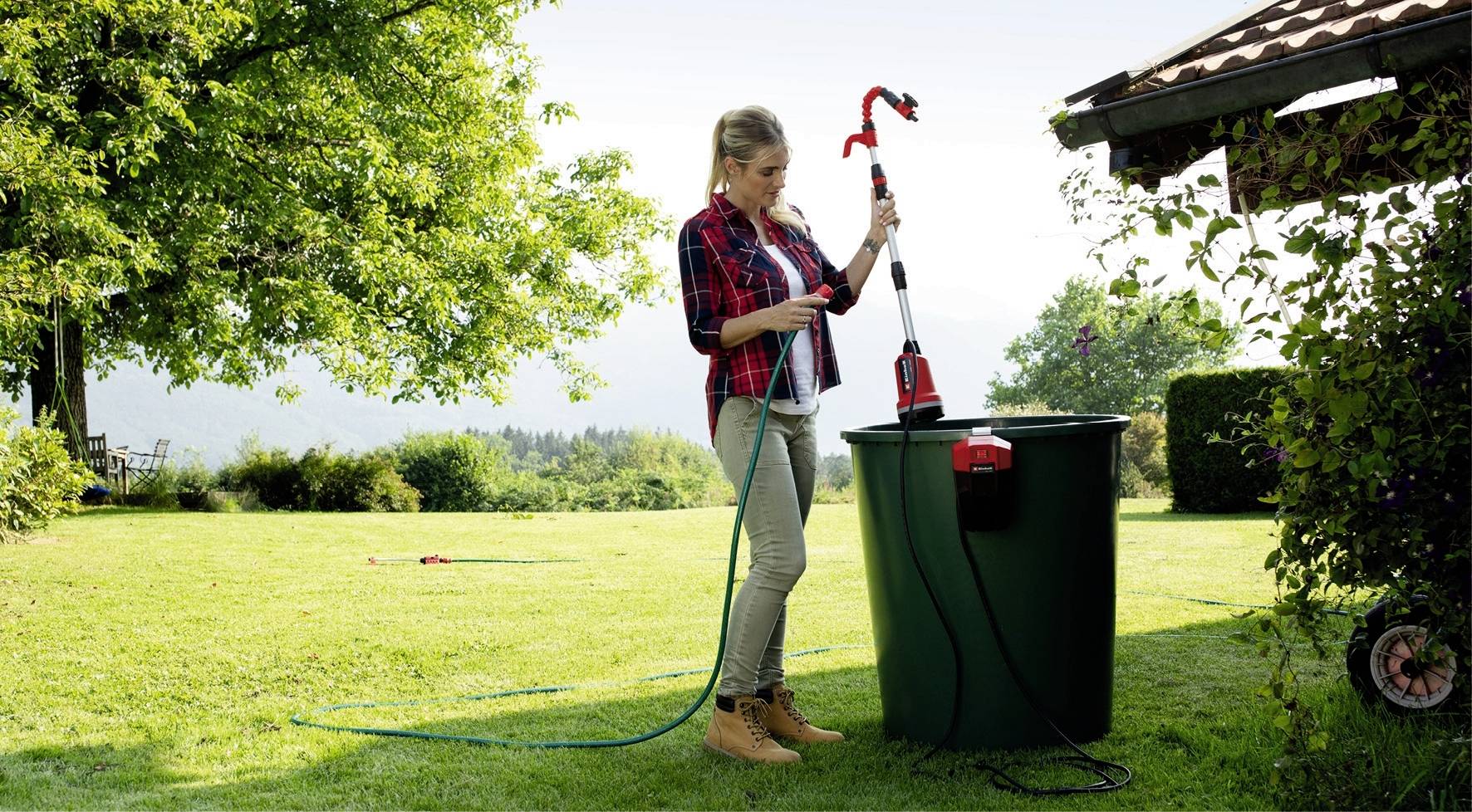 A woman is standing in the garden next to a large water tank and is holding a pump. She is placing the hose into the tank to pump water.