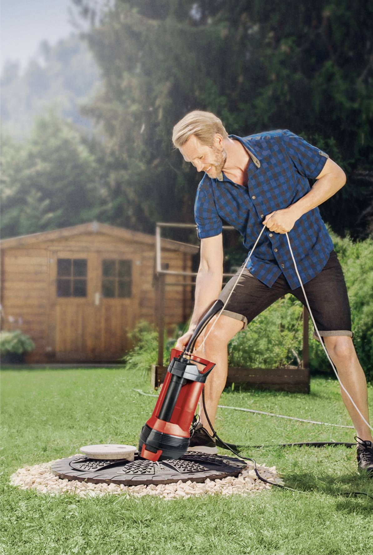A man in casual clothing is using a red water pump in a garden. A wooden shed and green vegetation are visible in the background.
