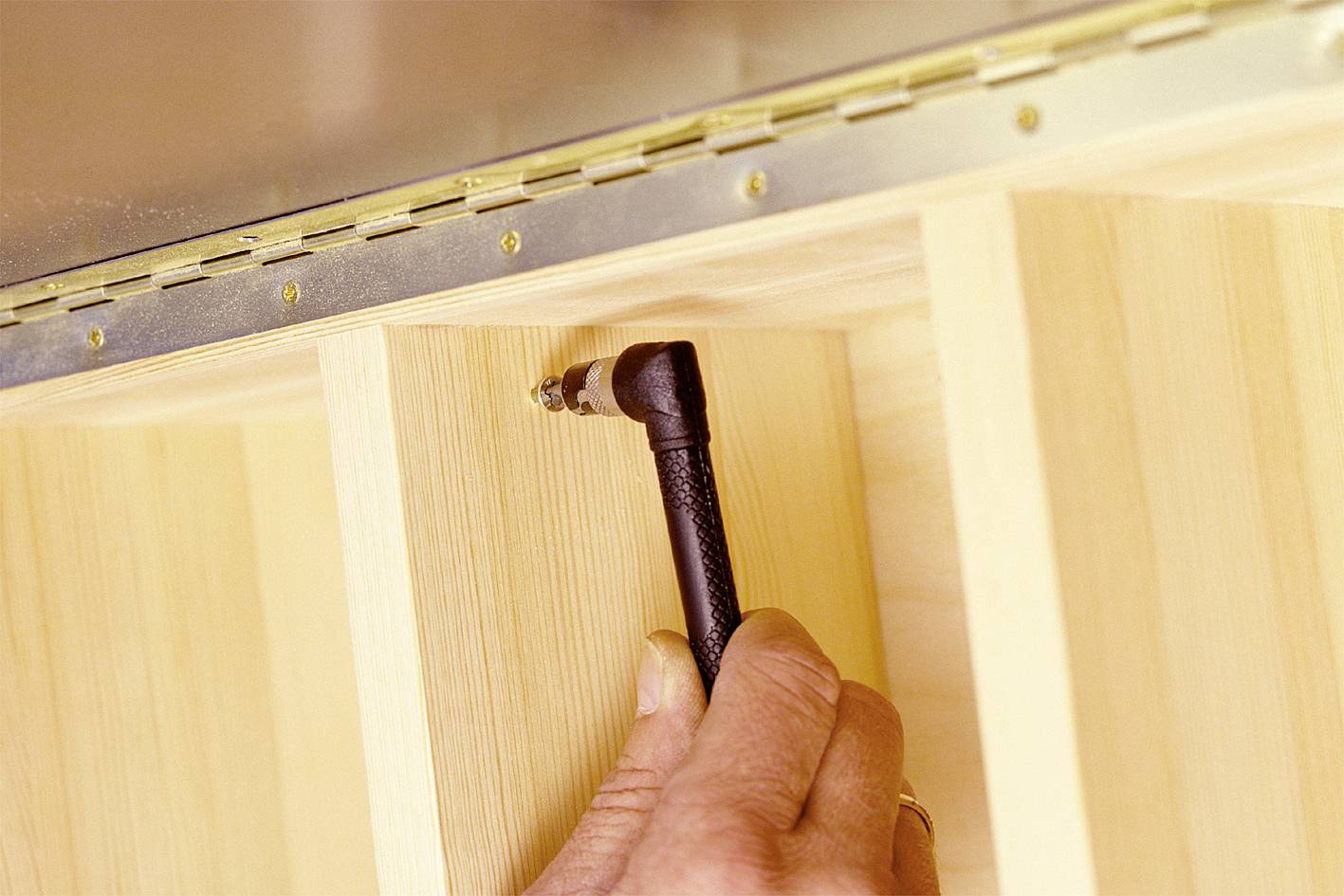 'A hand holds an Allen key, which is positioned at the top shelf bracket. A metal hinge is mounted on the shelf.'