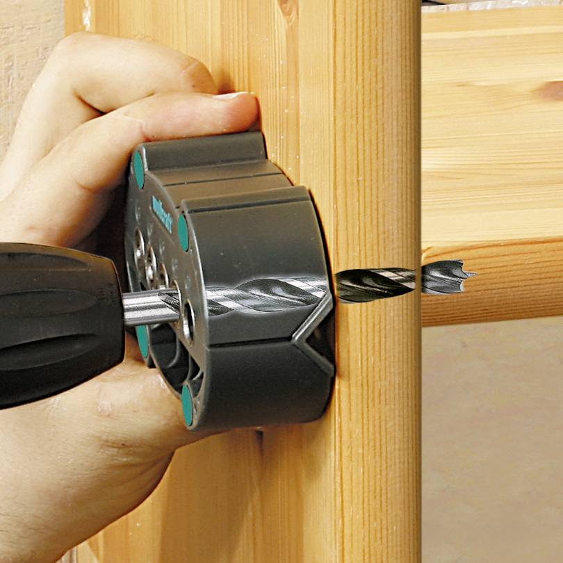 A hand is holding a drill with a hole saw attachment, drilling a hole in a wooden board.