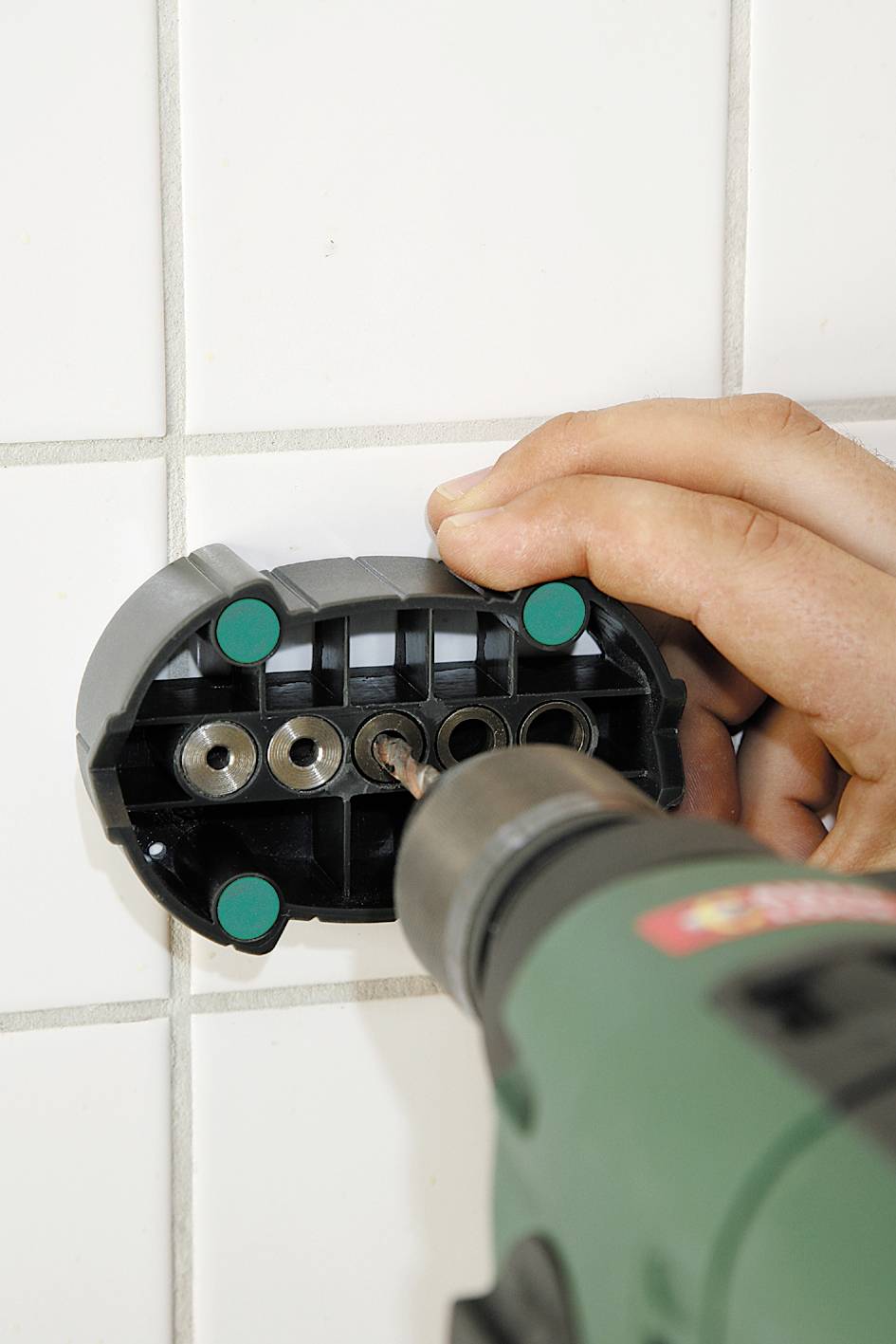 A hand is holding a drill mounting a bracket onto white tiles. The bracket has multiple fixing holes.