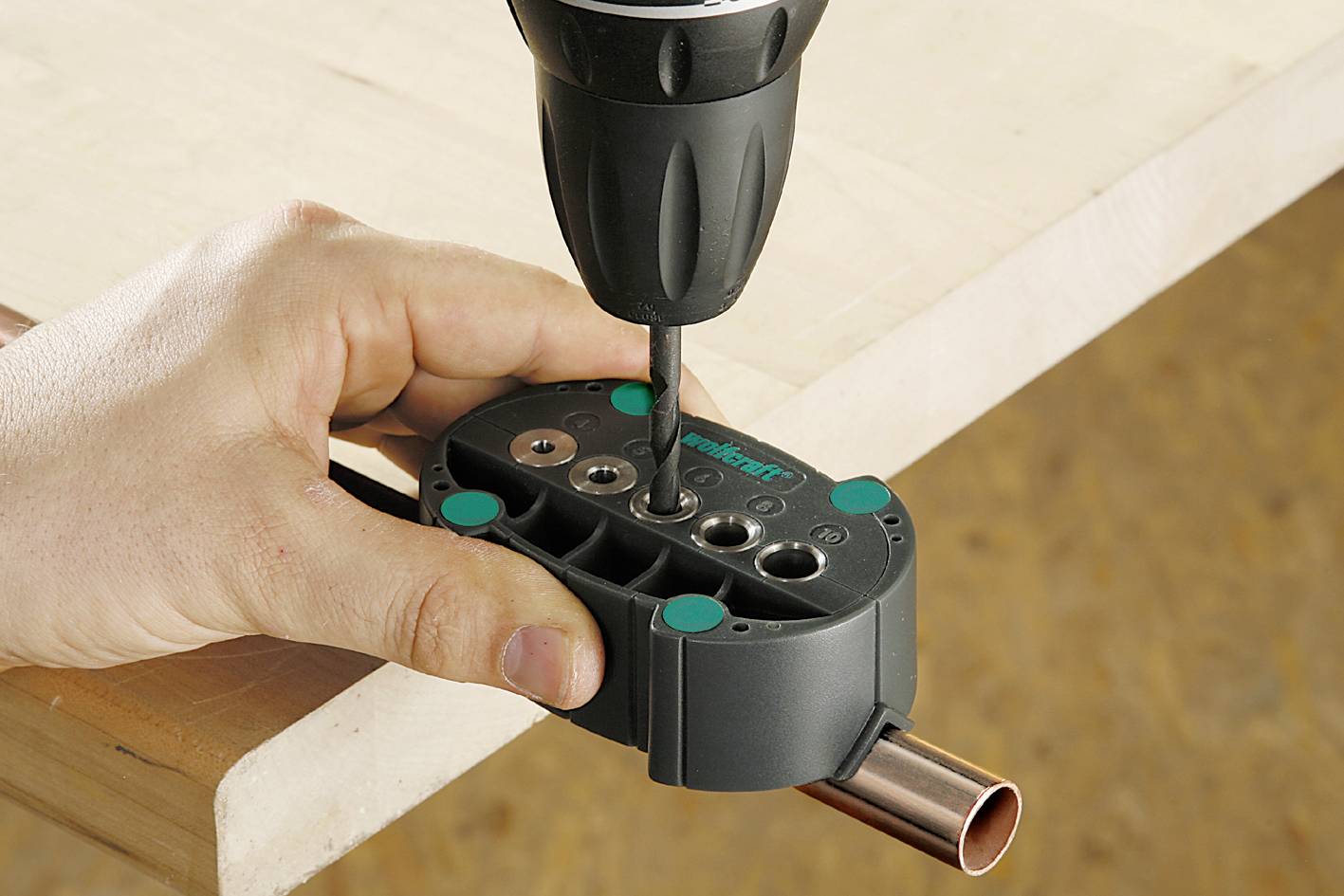 A hand holds a pipe on a table while a drill creates holes in a black plastic tool that surrounds the pipe.