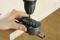 A hand holds a pipe on a table while a drill creates holes in a black plastic tool that surrounds the pipe.