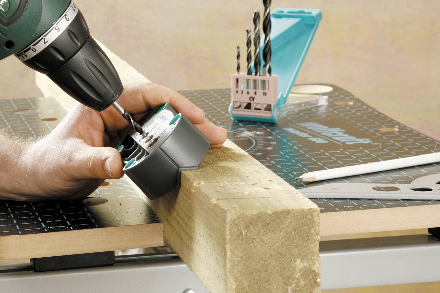 A person is drilling holes into a wooden block using a green drill, with a box of drill bits and a pencil lying nearby.