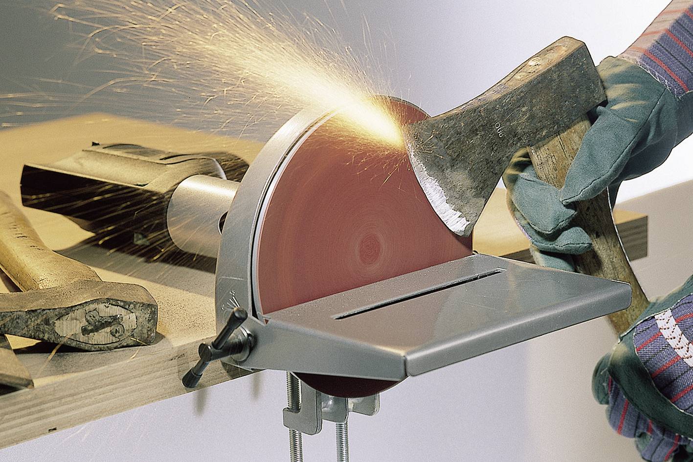 'A person is sharpening an axe on a grinding machine, with sparks flying. Other axes are lying on a wooden workbench.'