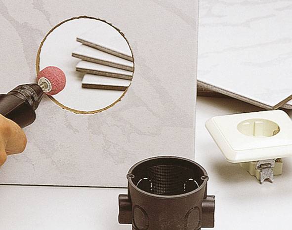 A tradesman is grinding a circular hole in a tile using an abrasive tool. Installation materials are laid out in the foreground.