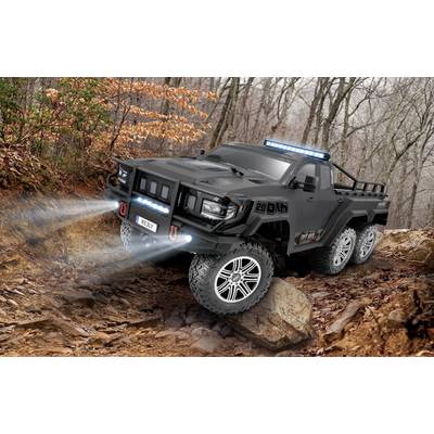 A black, six-wheeled off-road vehicle with gleaming headlights navigates a rocky trail lined with trees in the forest.