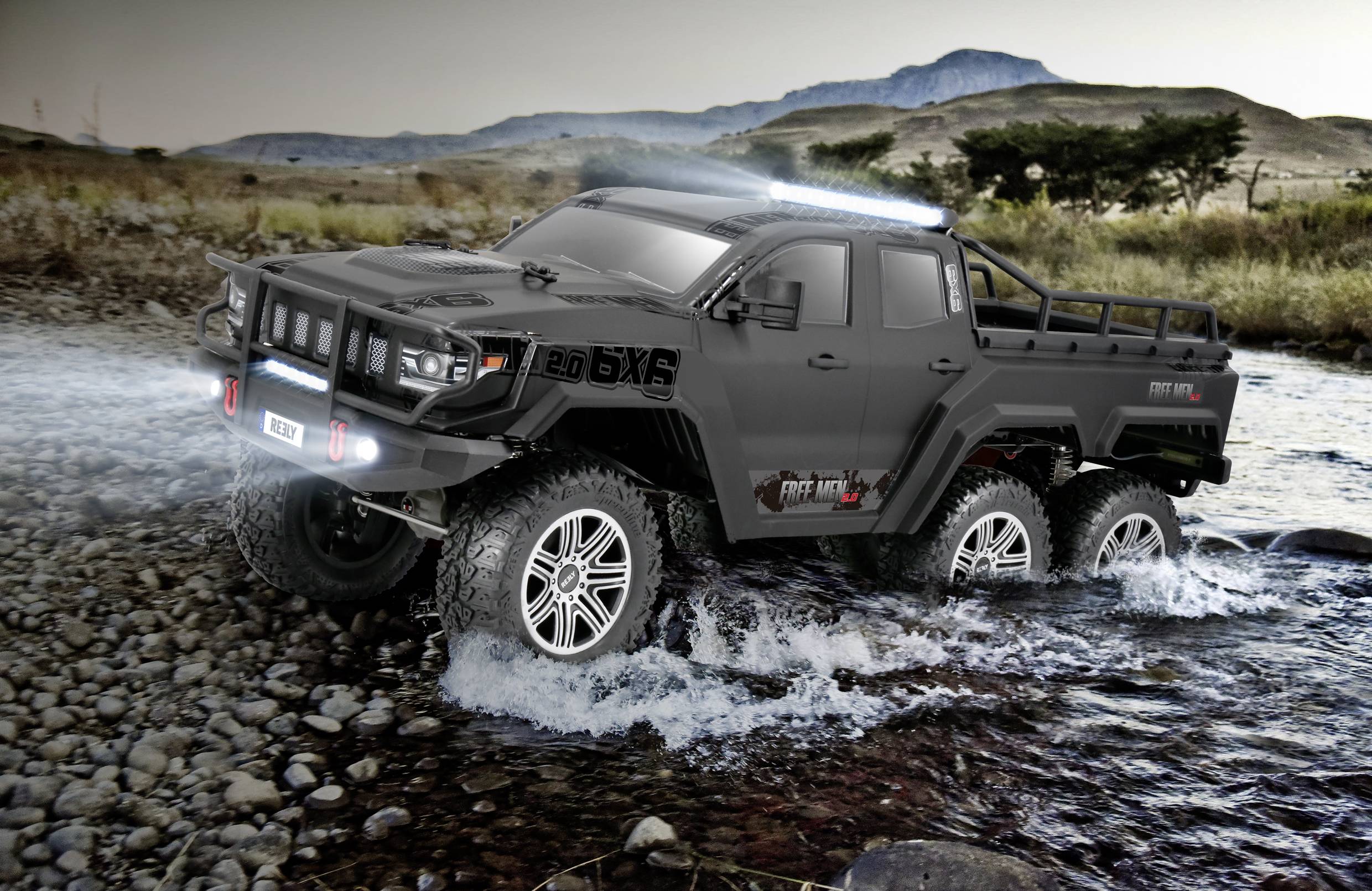 A black, six-wheeled off-road vehicle drives through a shallow stream in a rocky, rural landscape with hills in the background.