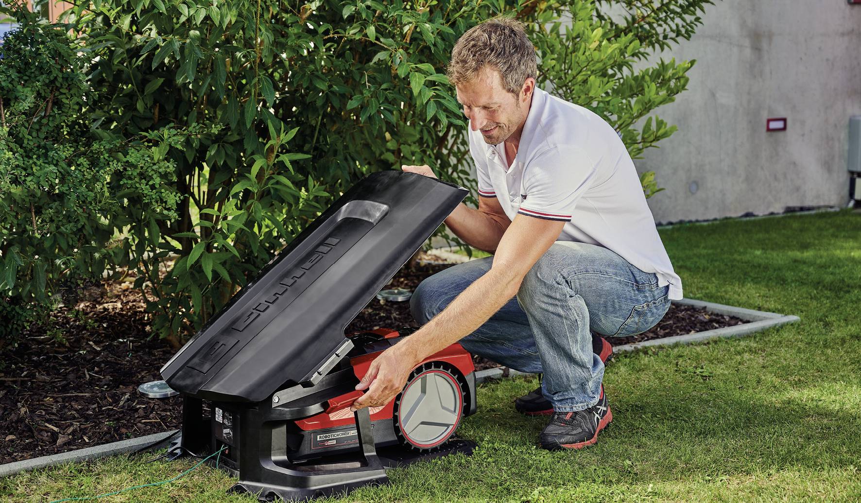 A man kneels in the garden and lifts the cover of a robotic lawnmower. It is daytime, and the garden is green and well-maintained.