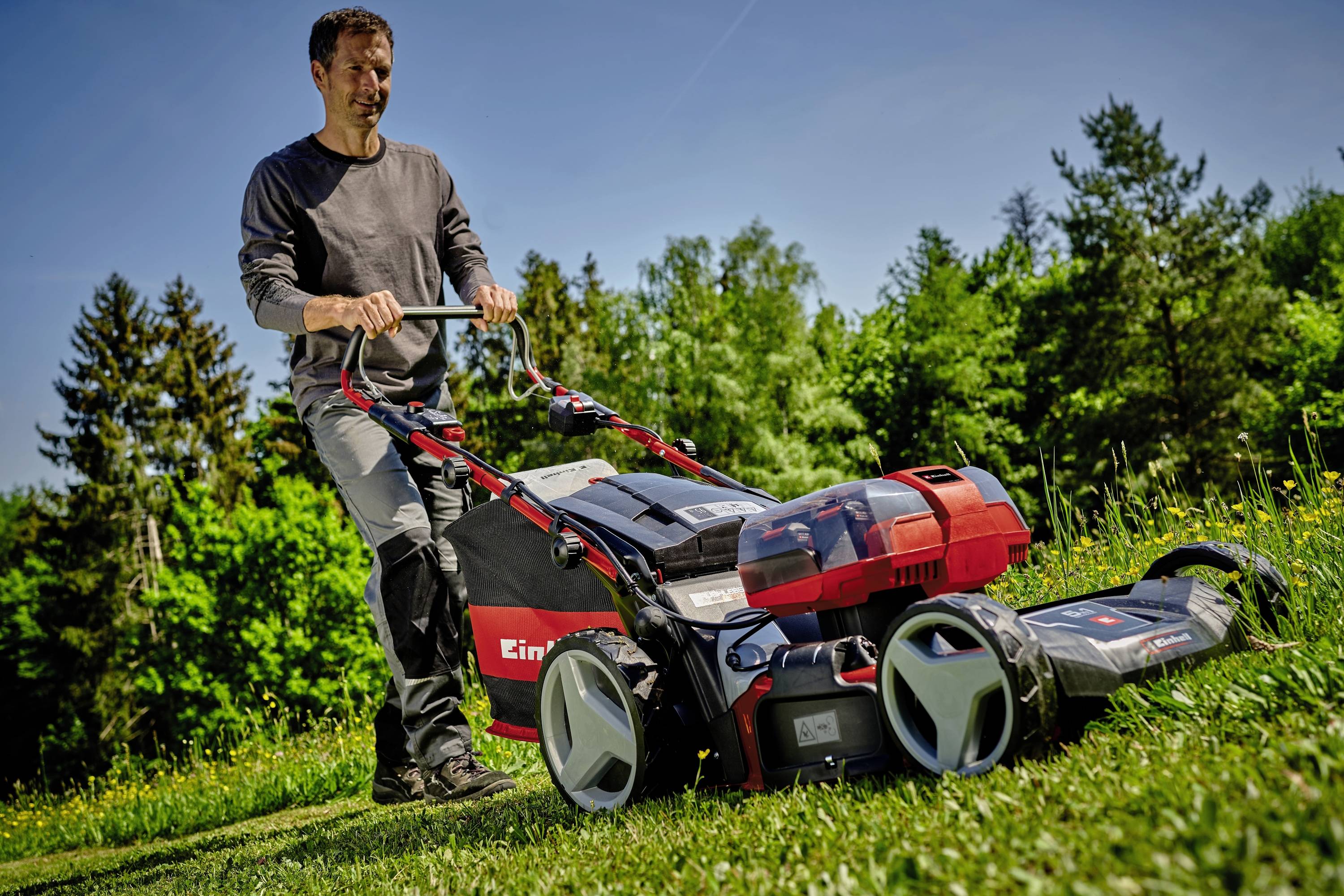A person is mowing the lawn with a lawnmower on a green lawn. Trees and a blue sky can be seen in the background.