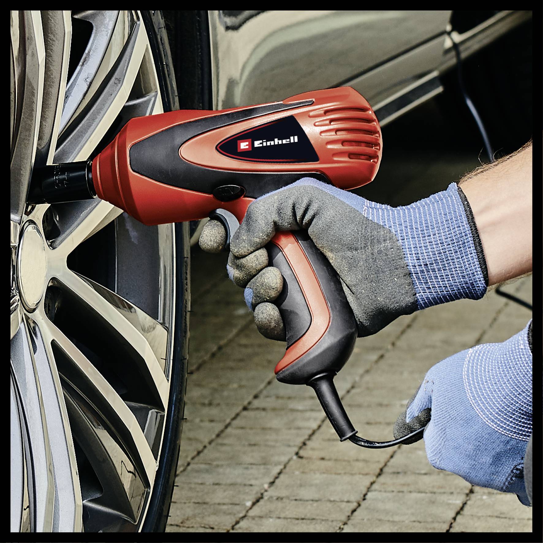 A person wearing blue gloves is using an electric impact wrench to remove a car tyre. The car is parked on a paved surface.