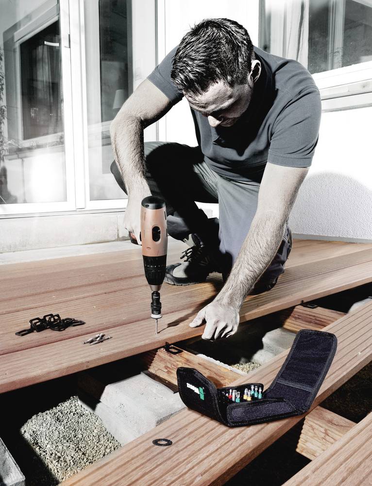A man is building a wooden terrace, kneeling with an electric tool. A tool bag with bits in the foreground. House facade in the background.