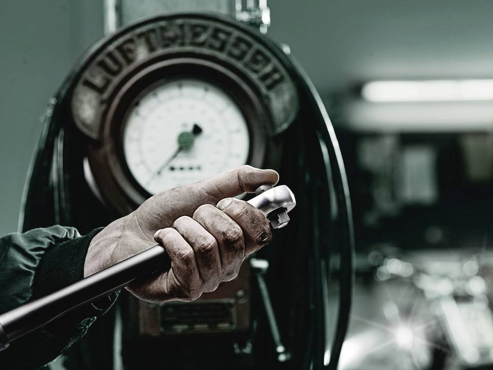 A hand is holding a spanner in front of an old 'air metre' device with a dial in a workshop. Background is out of focus.