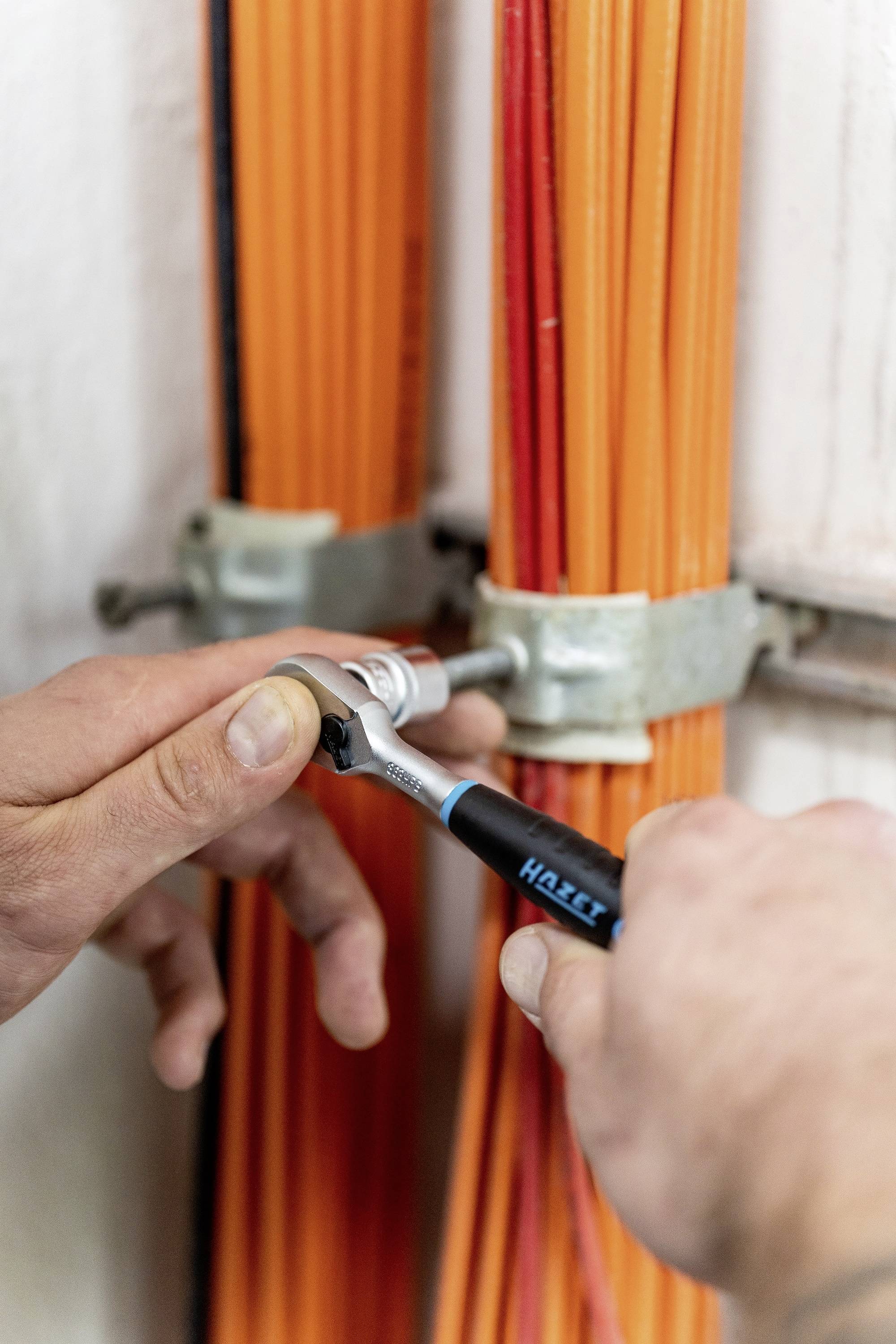 A person is tightening screws with a spanner to secure orange cable bundles to a wall mounting bracket.