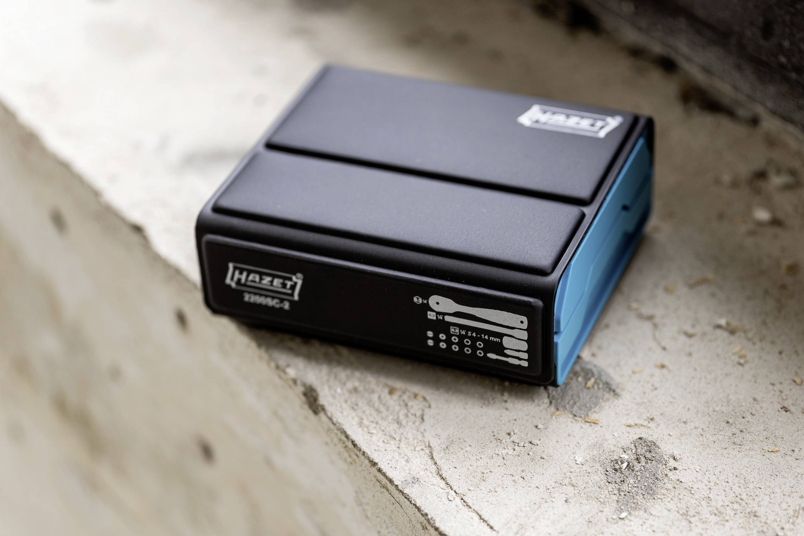 Black Hazet toolbox with blue side, placed on a concrete surface. Displays symbols for included tools.