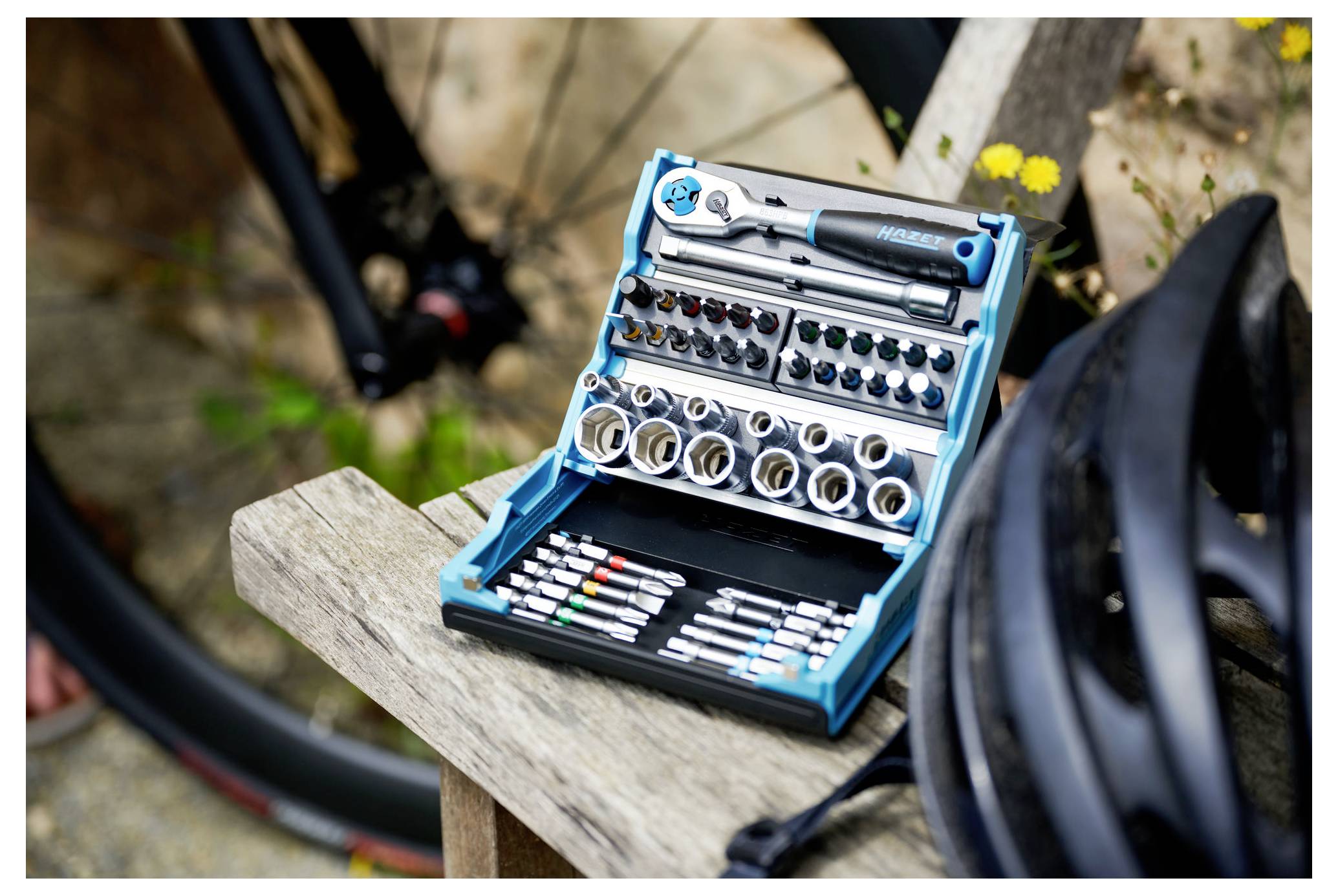 A toolkit with various hex keys and screwdrivers is open on a wooden bench beside a bicycle wheel and a helmet outdoors.