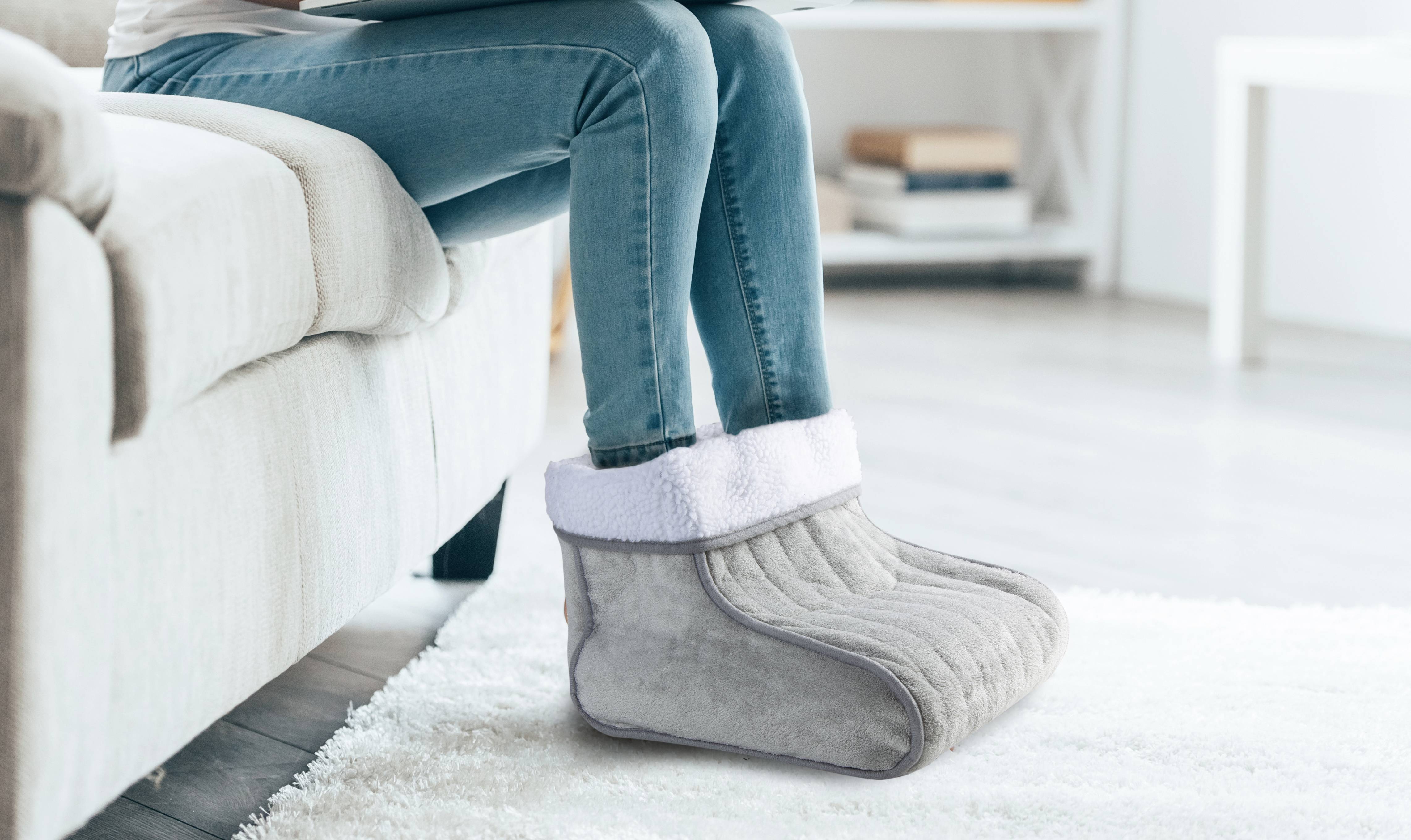 A person is sitting on a sofa and using a heated footwarmer in grey, with a living room background.