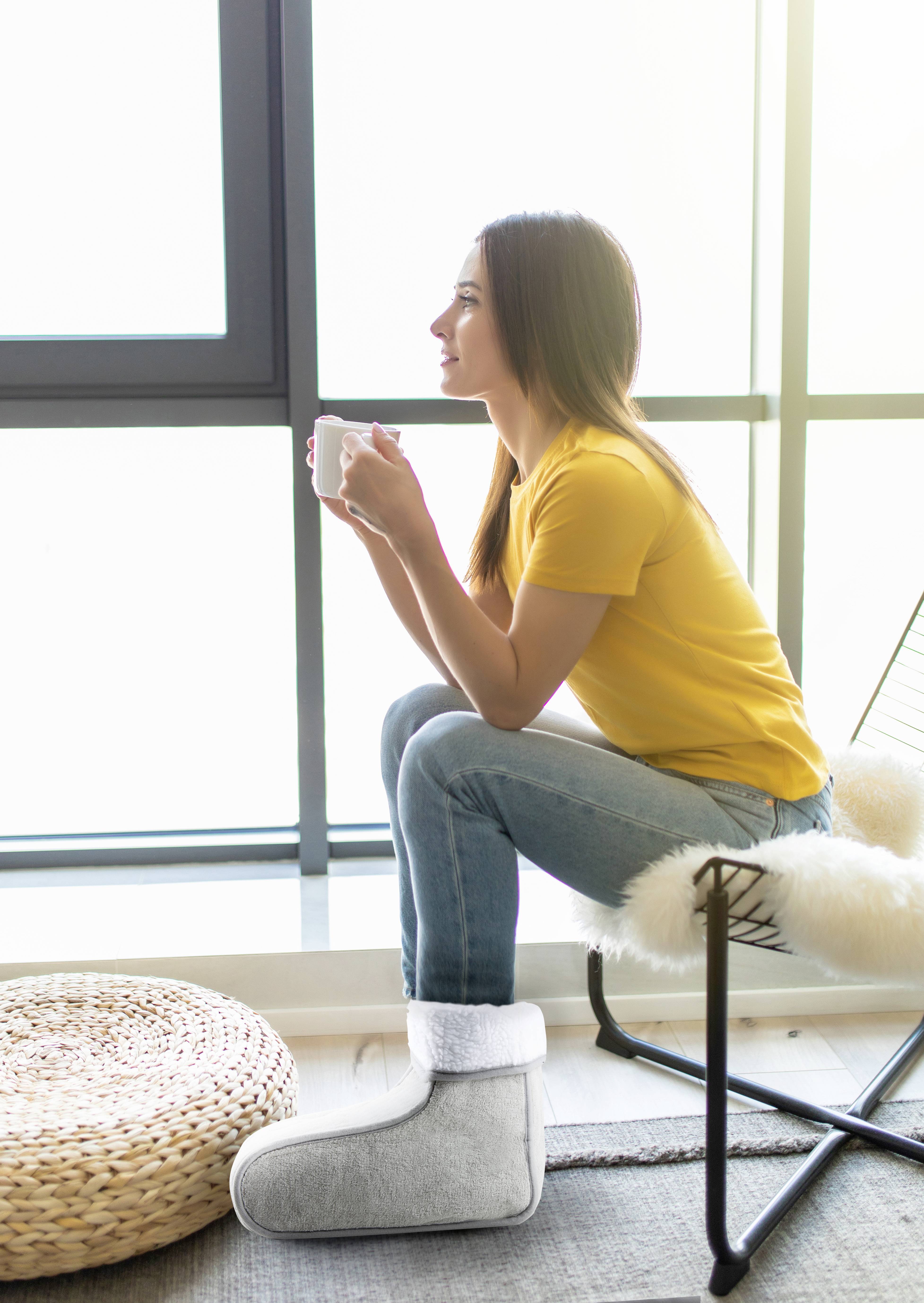 A smiling woman in a yellow T-shirt sits on a chair by the window, holding a mug, surrounded by bright daylight.