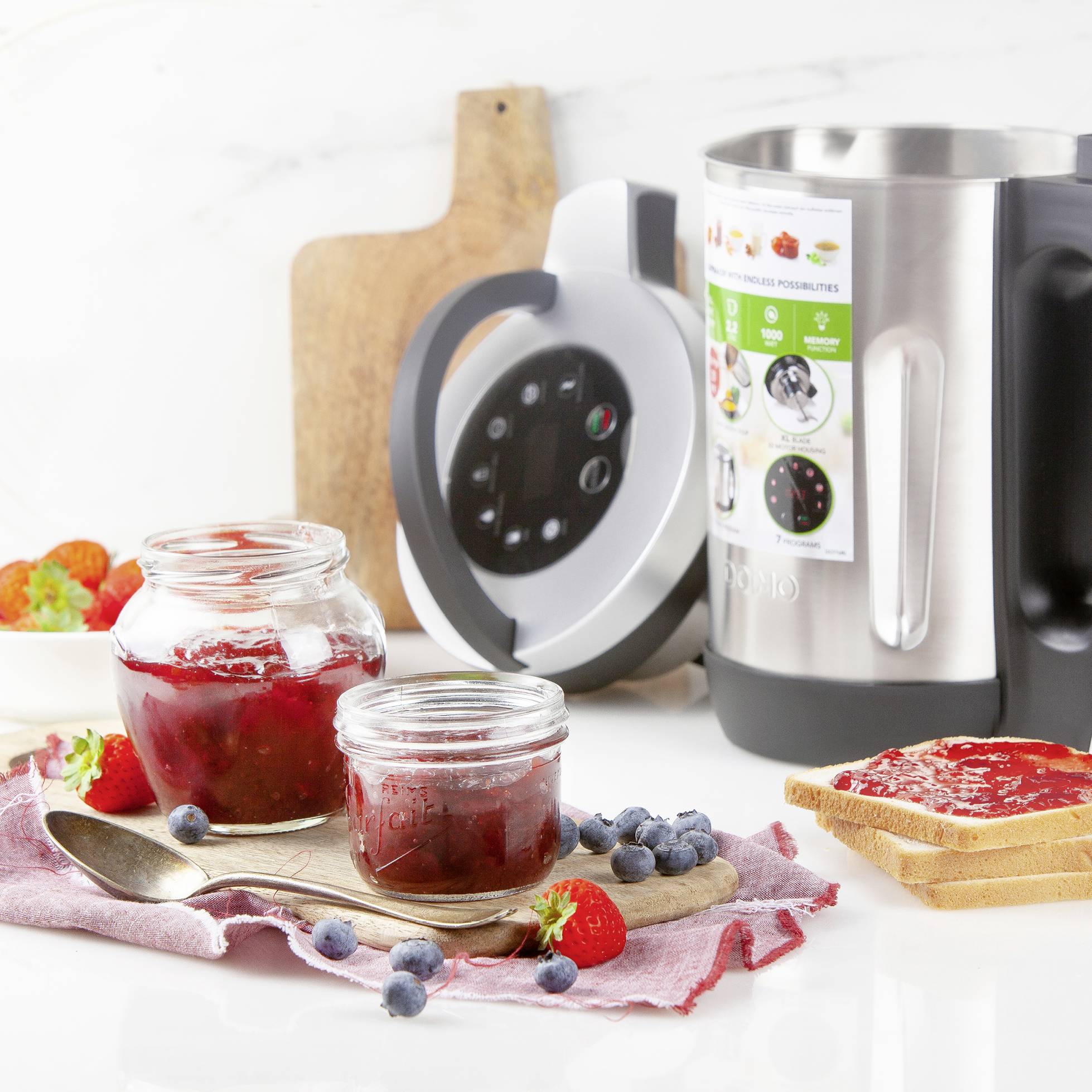 A blender with its lid removed stands next to a wooden board. In front of it are jam jars and fresh berries on a cloth.