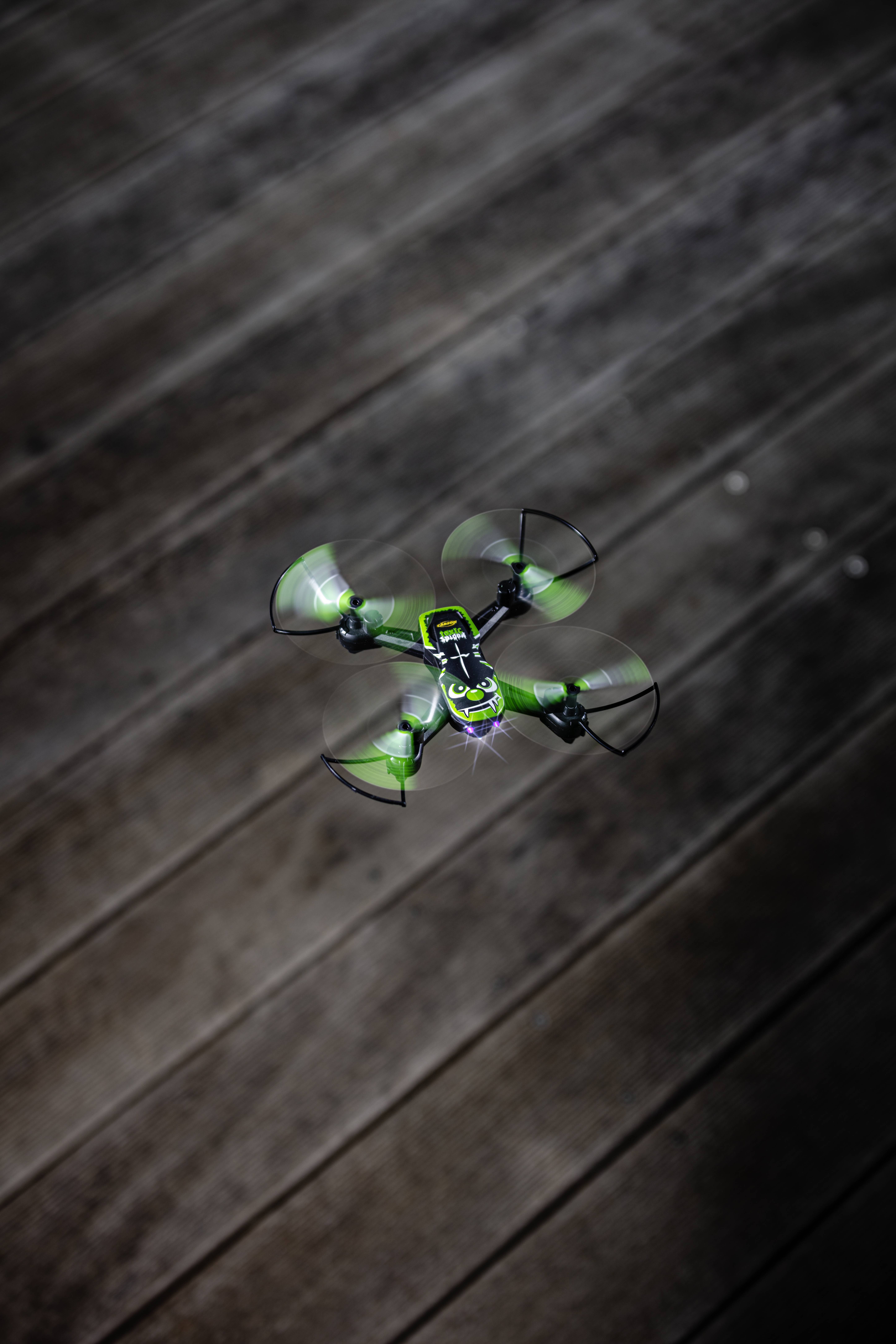 A green drone hovers above a wooden floor and is visible in flight.