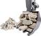 An excavator arm with a bucket is lifting a load of grey stones, with a small pile of stones lying on the ground in the background.