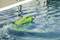 A green remote-controlled boat races swiftly across the surface of a swimming pool, splashing water and leaving waves in its wake.