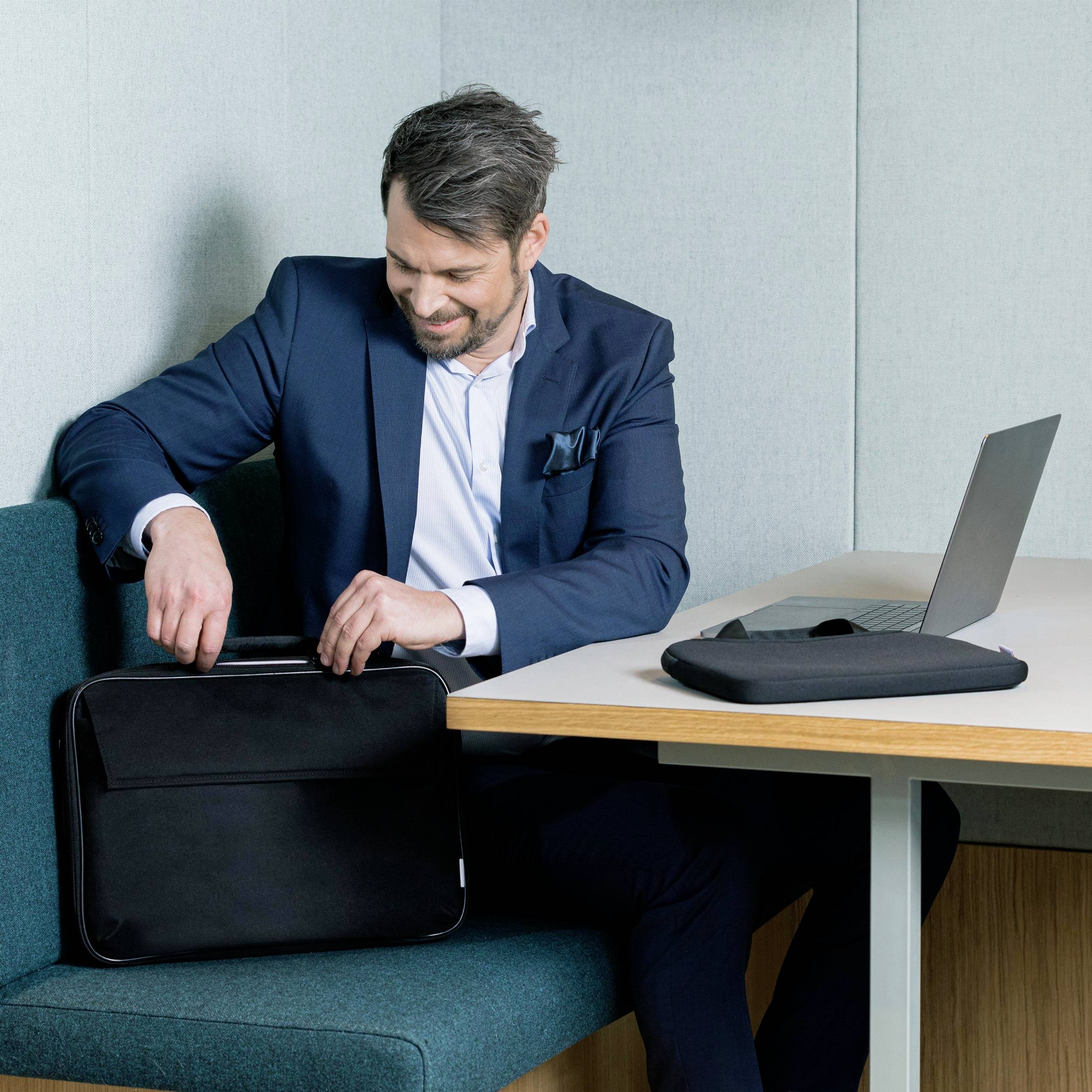 A man in a suit sits on a sofa in an office, holding a black briefcase and smiling at a laptop.
