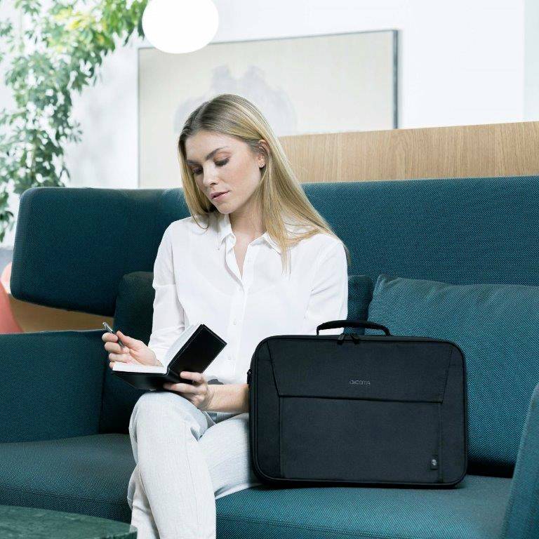 A woman is sitting on a sofa, writing in a notebook and holding a smartphone. A black laptop bag is lying beside her.
