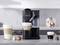 A black coffee machine is brewing coffee. Three cups with different coffee varieties are standing on a grey kitchen worktop.