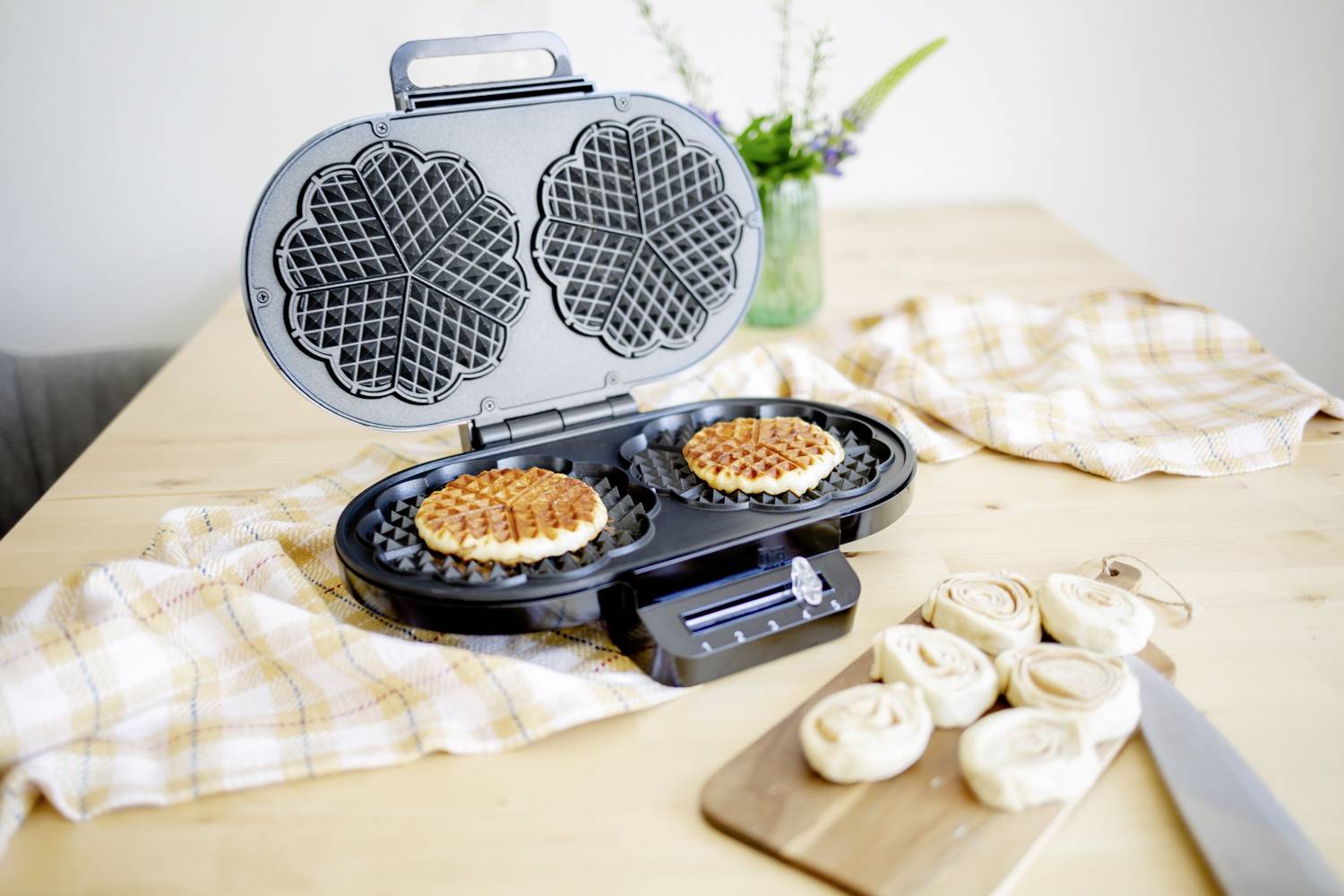 A waffle iron with two fresh waffles on a wooden table. Alongside are pieces of dough and a knife. In the background, a vase of flowers stands.