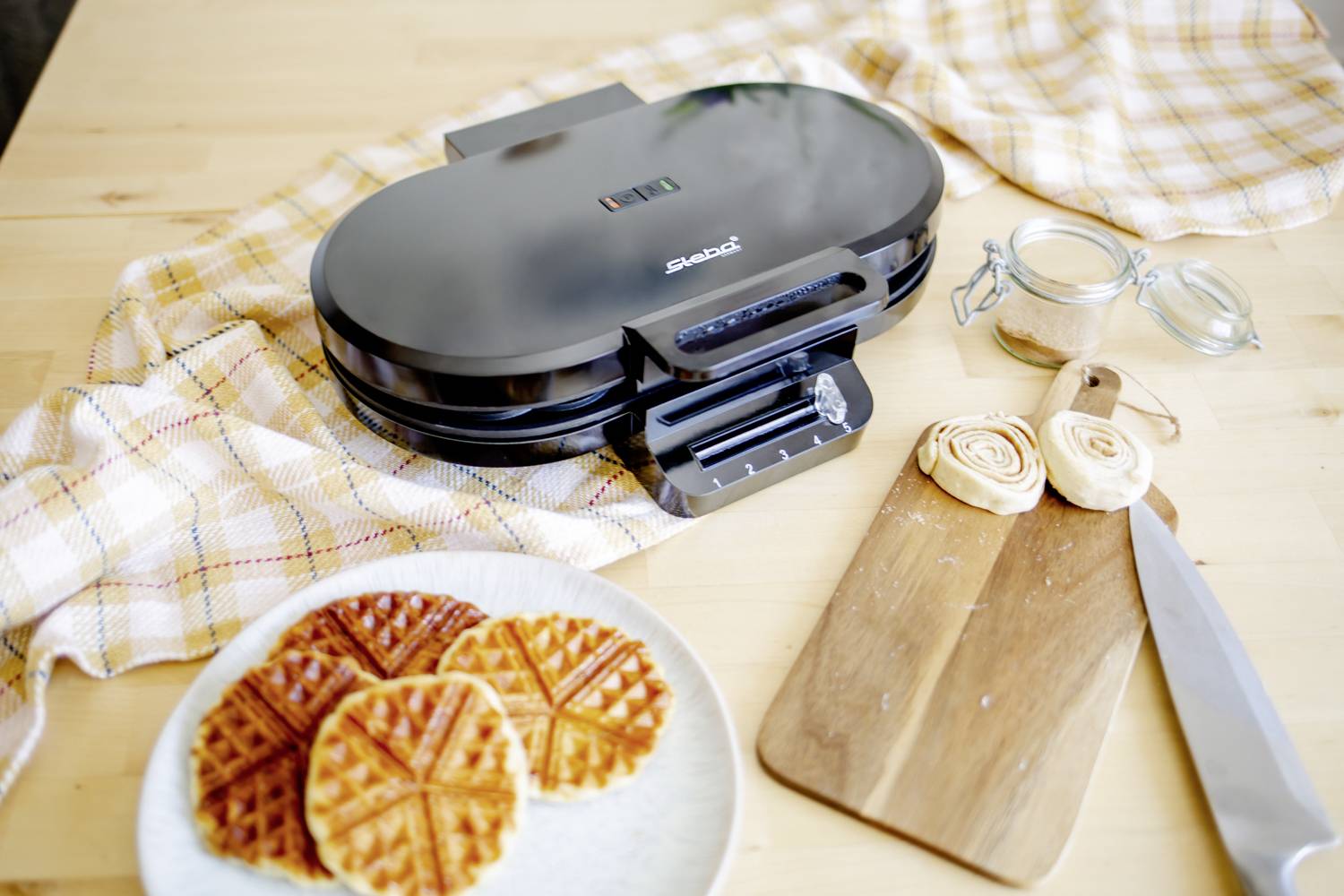 A waffle iron on a table with baked waffles on a plate, a wooden board with batter discs and a knife beside it.
