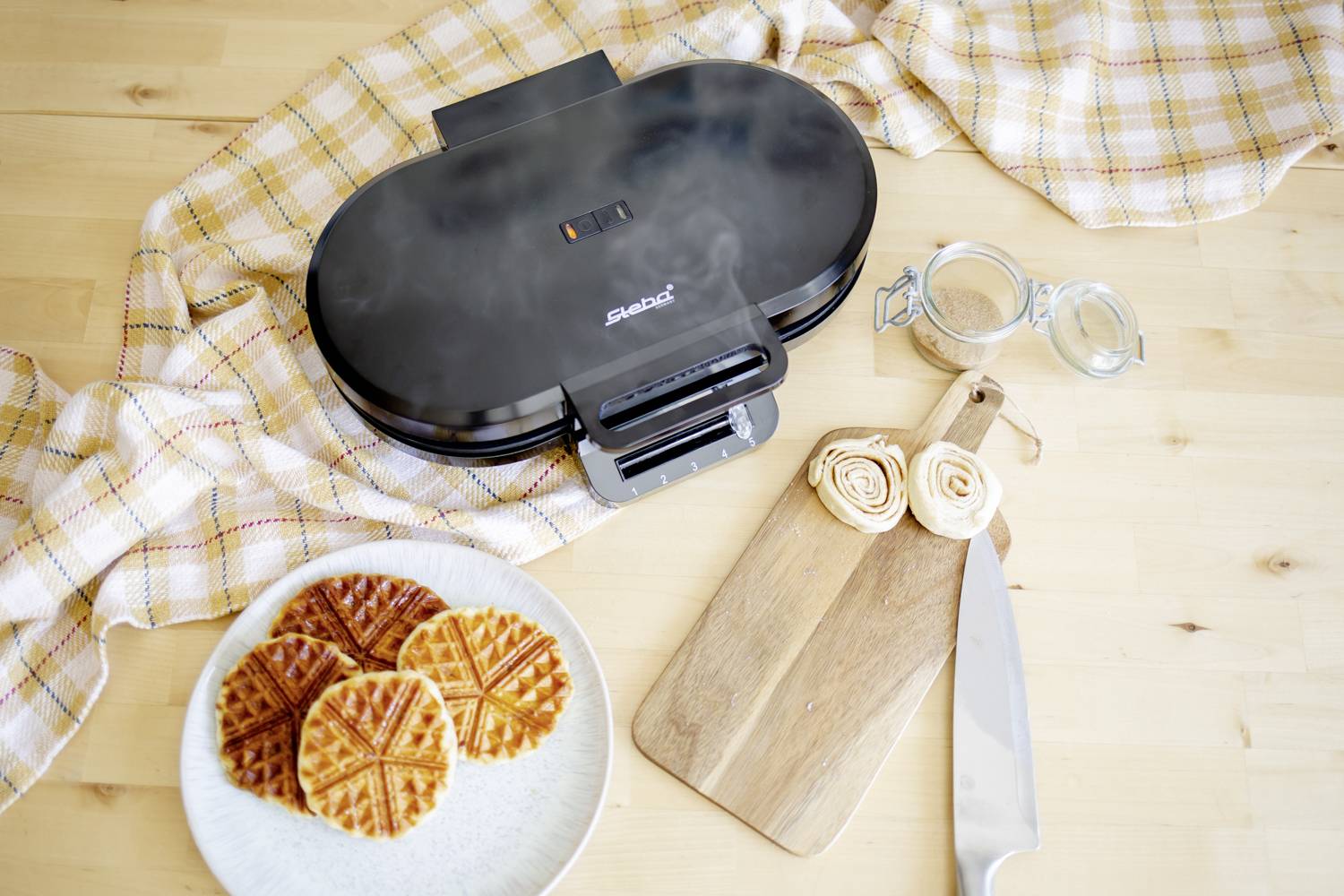 Waffle iron on a wooden table with brown waffles on a plate. Alongside are a chopping board, a knife, and a glass lid.