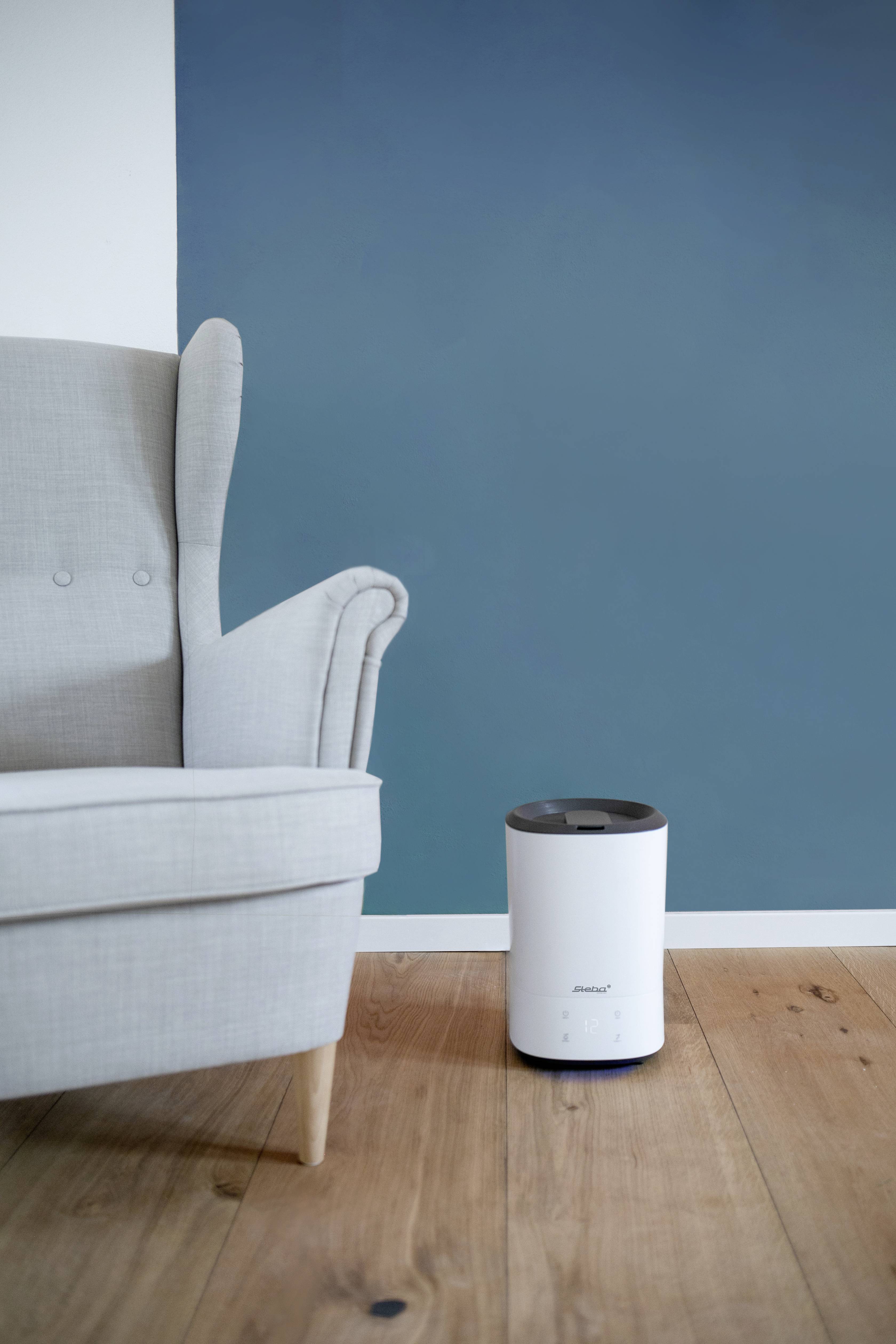 A white air humidifier stands on a wooden floor next to a light grey armchair in front of a blue wall.