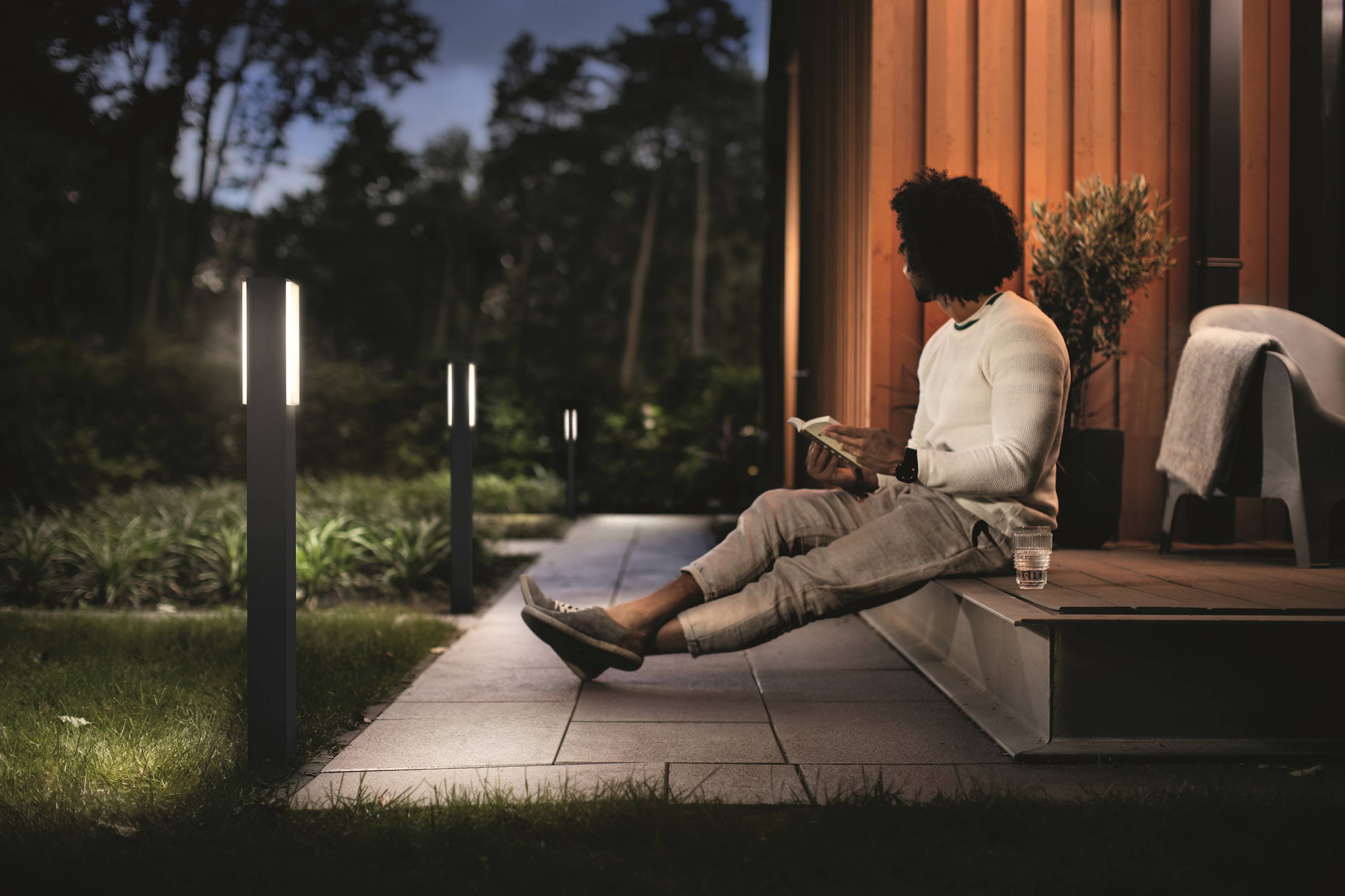 A man sits on a terrace reading at dusk. Modern garden lighting illuminates the path, and surrounding trees are visible.