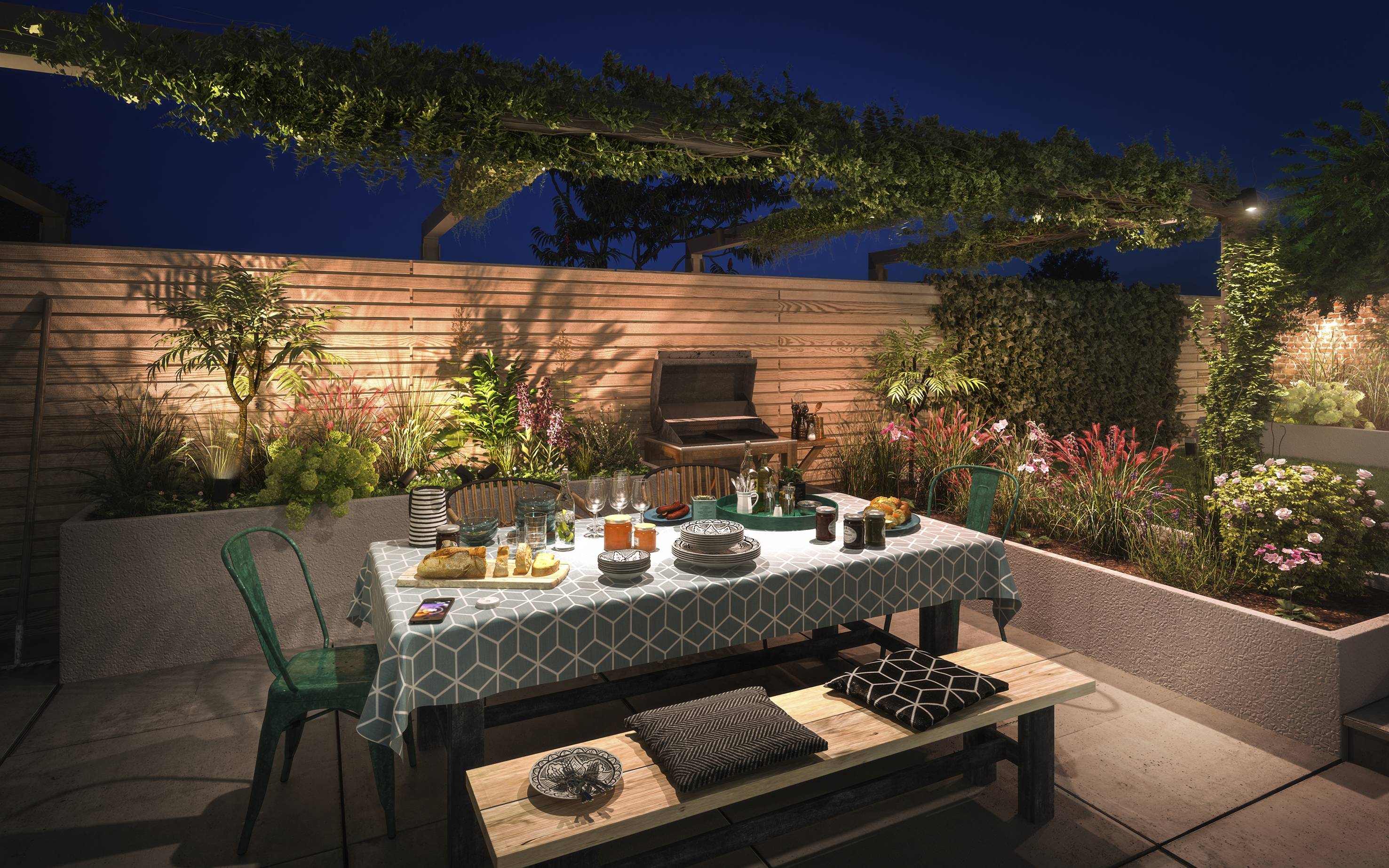 An outdoor area at night with a laid table for an evening meal. Plants and a wooden partition are in the background.