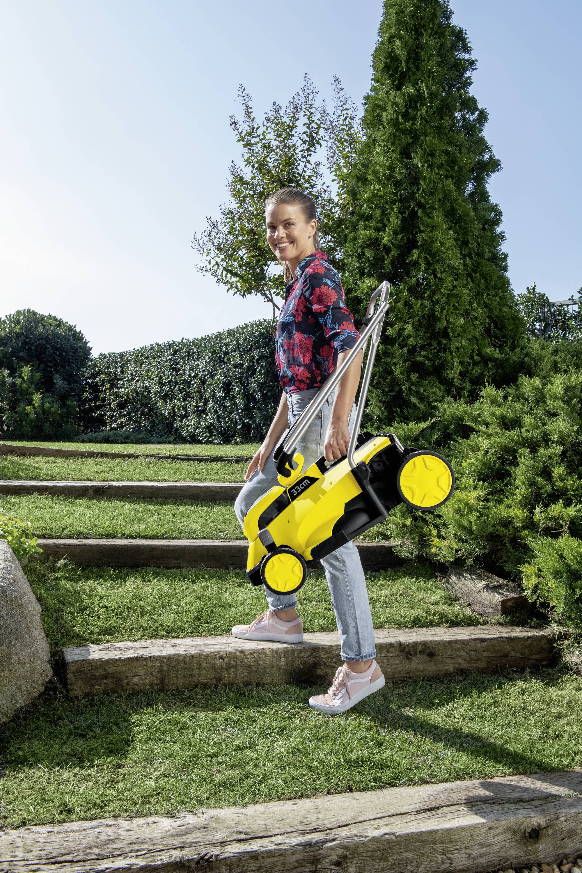 A woman is carrying a yellow, lightweight lawnmower up a grassy stone staircase. Shrubs and a tree are visible in the background.