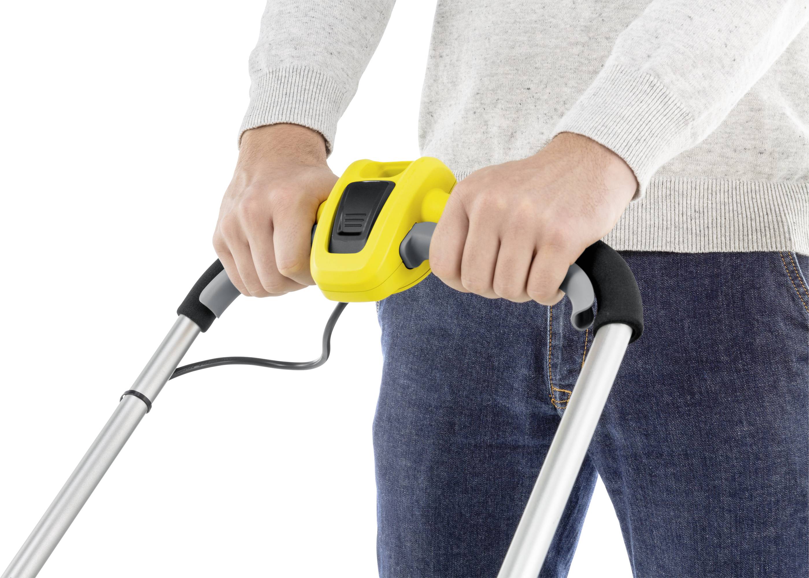 A person is holding the handles of an electric lawnmower with a yellow control panel. They are wearing a grey jumper and blue jeans.