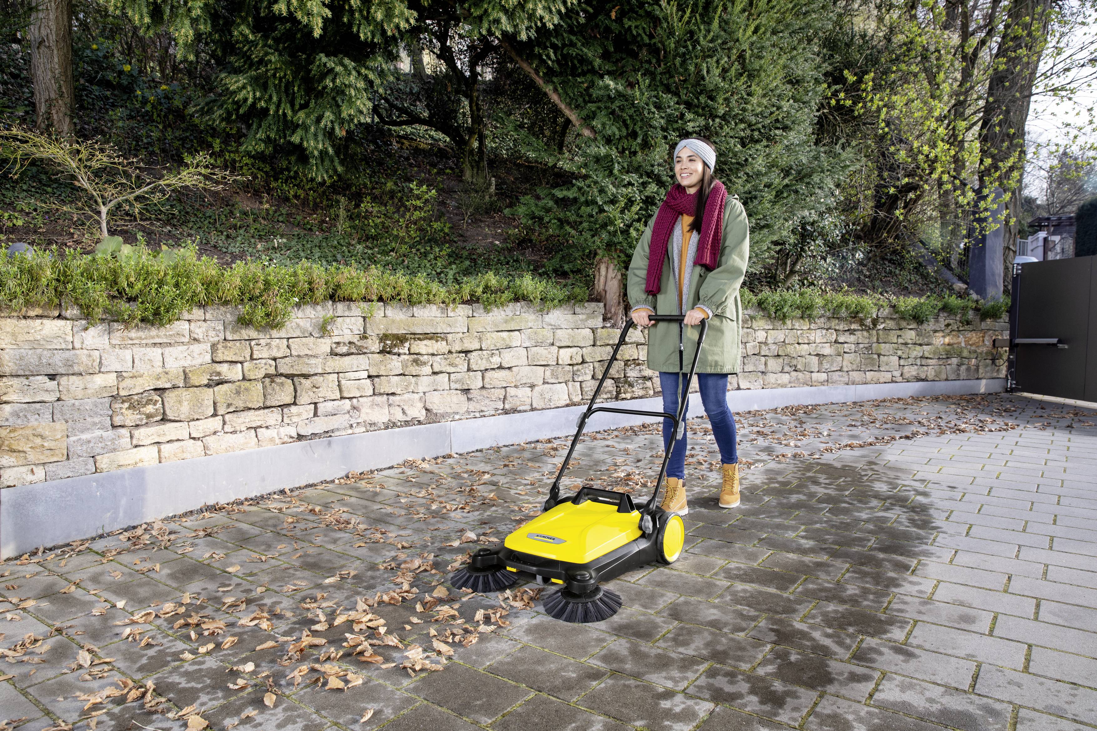 A person is sweeping with a yellow street sweeper on a pavement covered with leaves. A stone wall is visible in the background.